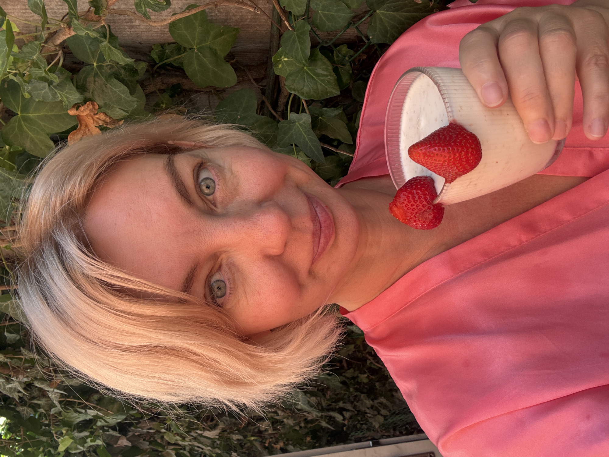 A blonde woman with fair skin and blue eyes, wearing a pink silk robe, holds a glass of a light-colored beverage topped with two strawberries. She is outdoors, with green ivy and a wooden fence in the background. The lighting suggests it is daytime. The image is a selfie. The scene is not identifiable as being in Gelendzhik, Russia.