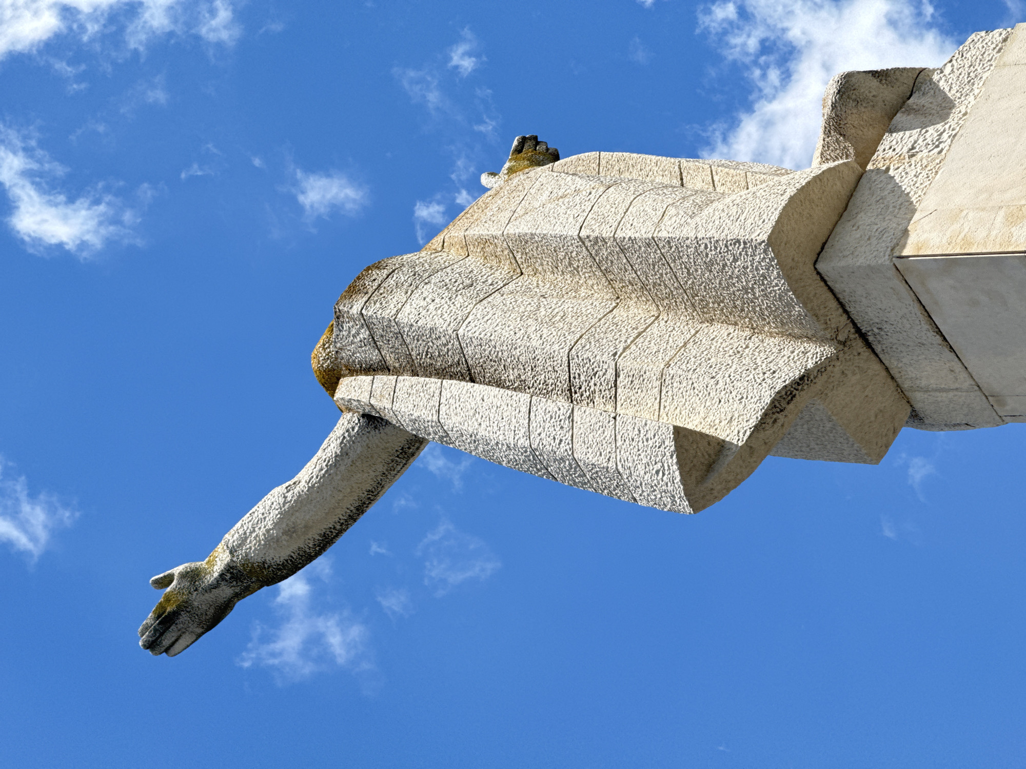 The image shows a close-up, low-angle view of a large, stone statue against a bright blue sky with scattered white clouds. The statue is abstract and geometric, with textured, light-colored stone. The visible portion includes a raised arm outstretched to the left, with fingers splayed. The body of the statue is blocky and angular, with a draped appearance. There is some green moss or lichen on the upper parts of the statue. The base of the statue is also visible, resting on a darker stone pedestal. This statue is part of the Monument to the Bulgarian Soviet Friendship, located on a hill in the city of Shumen, Bulgaria.