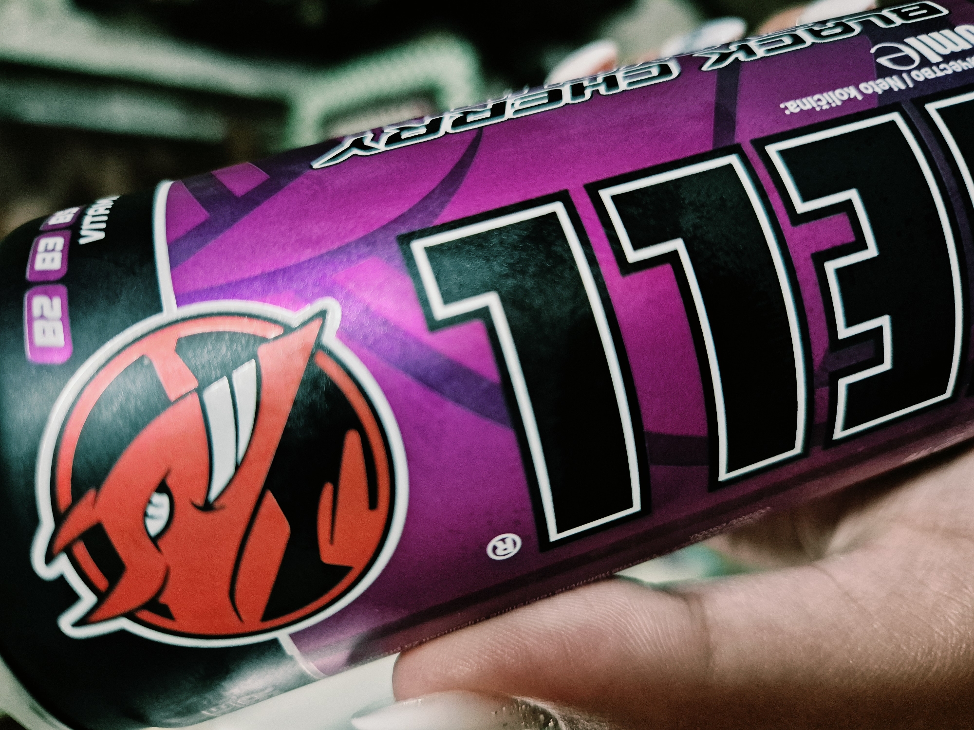 A purple and black energy drink can, identified as "HELL" brand, is held in a left hand in the city of Iskrets, Bulgaria. The can features a red devil-like character logo with horns and prominent teeth within a circular design. The words "B2 B3 VITAM" are visible near the top. The flavor "BLACK CHERRY" is printed vertically on the right side of the can, with partially visible text "Umie" and "Neto kolicina:" below it, indicating net quantity. The hand exhibits light skin tone and polished fingernails. The background is indistinct and out of focus.