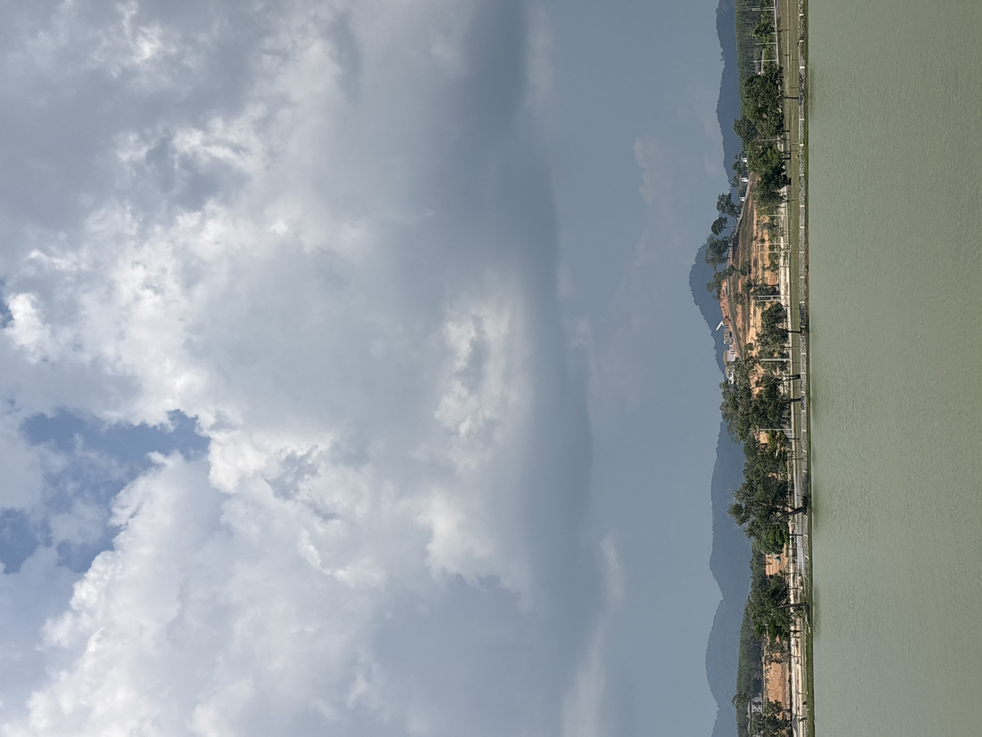 A wide landscape shot captures a serene scene comprising a vast sky, a developed shoreline, distant mountains, and a body of water, viewed under a partly cloudy daytime sky. The horizon line runs horizontally across the middle of the frame, separating the sky from the terrestrial and aquatic elements.
The upper half of the image is dominated by the sky, filled with a mixture of bright, fluffy white clouds and more expansive, darker grey clouds, indicating variable weather. Sections of clear blue sky are visible, especially towards the upper left.
Below the sky, a strip of land forms the shoreline, stretching across the entire width of the image. This land shows signs of human activity, with areas of reddish-brown earth suggesting recent clearing or construction. A variety of green trees, both mature and young, are interspersed along the water's edge and extend inland up gentle slopes. Some structures, resembling buildings or houses, are discernible among the trees and on higher ground. A path or road appears to run parallel to the water's edge.
In the background, a range of hazy, bluish-grey mountains with undulating peaks rises, their contours softened by atmospheric perspective, adding depth to the scene.
The lower portion of the image features a calm body of water, appearing a muted greenish-grey color, with only slight ripples disturbing its smooth surface. The lighting suggests it is daytime, possibly late morning or early afternoon. The landscape's features, including the vegetation and mountain formations, align with the provided context of Choeng Thale, Thailand. No specific human activity is overtly visible, nor is any text present in the image.