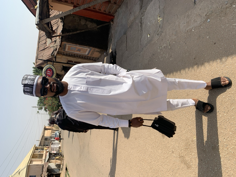 The image depicts an outdoor street scene in the city of Jos, Nigeria, under clear daylight conditions.
In the foreground, centered, an adult male stands facing the camera. He wears a white long-sleeved tunic (kaftan style) with a mandarin collar and a short placket, paired with matching white trousers. His head is adorned with a black and white patterned cap, and he wears dark sunglasses. A short beard is visible. He holds a small, structured black bag with a top handle in his left hand, while his right hand rests in his trouser pocket. Black open-toed sandals are on his feet, and a beaded bracelet is on his left wrist.
In the left background, a second adult male, partially visible, walks away from the camera, dressed in dark clothing. Further down the street on the left, a light-colored vehicle is partially visible, along with a distant, indistinct figure.
The environment consists of an unpaved or dirt-covered street, light brown in color, with visible cracks and an uneven surface. Strong shadows are cast on the ground from the right side of the frame, indicating an overhead sun. On the left side of the street, buildings with varied facades and roofs are visible; one structure is a two-story building with a light-colored roof, followed by simpler buildings with corrugated iron roofs. On the right side, closer to the camera, is a single-story building with a faded brown-orange exterior and a corrugated iron roof, featuring a circular red and yellow "33" Export beer sign. Adjacent to it, another structure with a red vertical support and a corrugated iron roof is present. Faded painted advertisements are visible on these background buildings. The sky is clear and light blue.