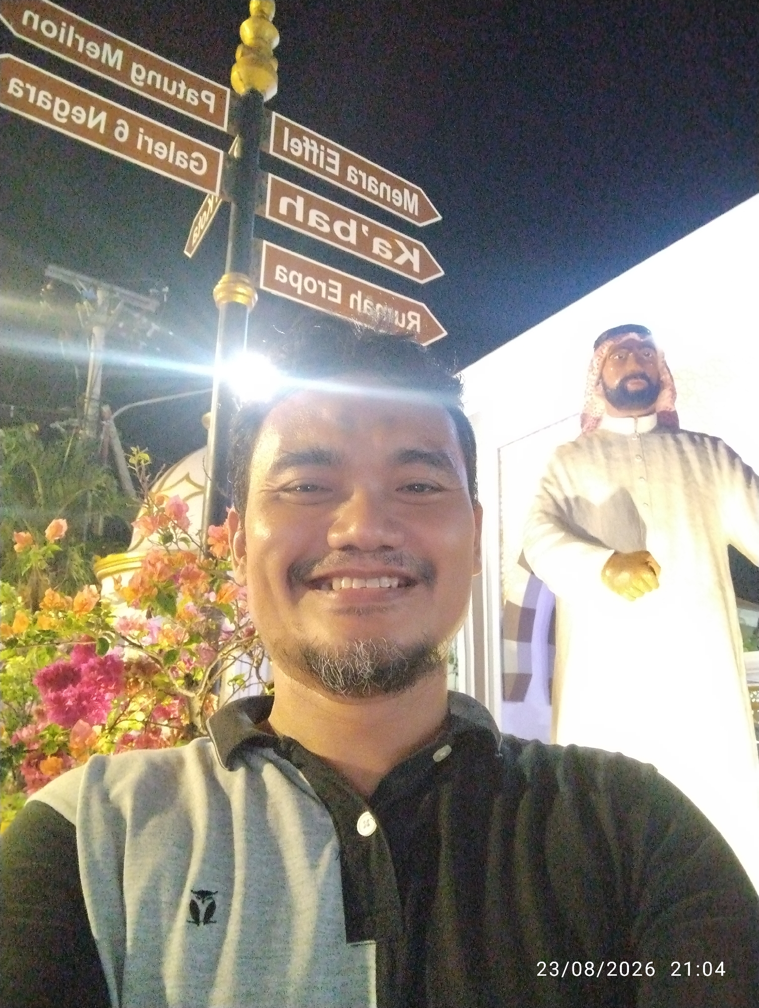A man is taking a selfie in front of a signpost and a statue in the city of Madiun, Indonesia. The signpost has several directional signs with text in both Indonesian and what appears to be Arabic. The man is smiling and has a goatee. Behind him, there is a statue of a man wearing traditional Middle Eastern clothing. The background shows some flowering plants and a bright light source, likely from artificial lighting. The date stamp on the image reads 23/08/2026 21:04, suggesting the photo was taken at night.