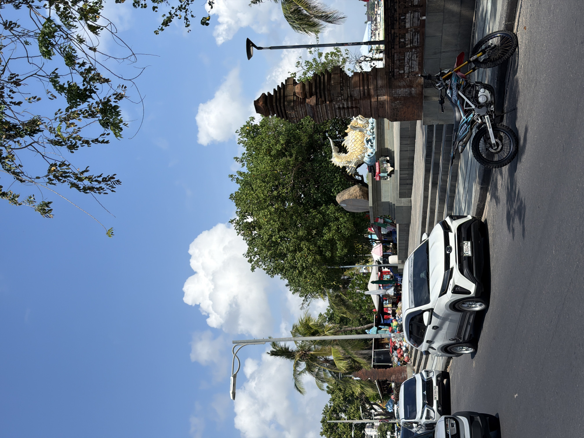 This is a vertical, eye-level shot of a street scene in Kuta, Indonesia, likely taken during the day. The sky is a bright blue with scattered white clouds. Lush green trees and palm trees line the street, some of which are in the foreground obscuring parts of the sky.
Dominating the mid-ground is a weathered brick temple-like structure with a tiered roof, adorned with a golden dragon statue. Adjacent to the structure, a modern dirt bike is parked on the paved road. Further down the street, a white SUV is parked, with its license plate visible: "AT6945". Another white vehicle, possibly an SUV, is parked behind it. The road is asphalt, and there are stone steps leading up to the temple structure.
On the right side of the frame, there's a glimpse of market stalls with colorful fabrics and various goods, suggesting a vibrant commercial area. The overall impression is of a lively tourist destination. The sun appears to be high in the sky, casting sharp shadows, indicating it's likely midday. No specific emotions are evident from the static image.