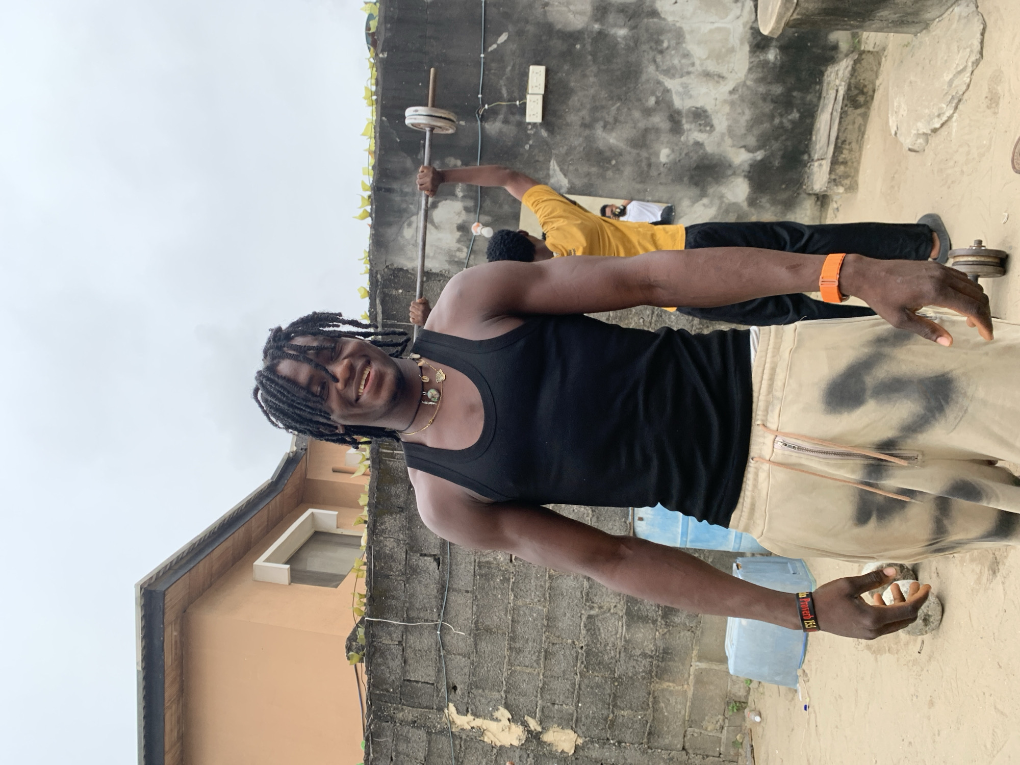 In Ado, Nigeria, a young Black male with braided hair smiles at the camera. He wears a black tank top, tan pants with black tie-dye markings, and a beaded necklace. On his left wrist is a black wristband with red text, and on his right is an orange wristband. He stands on a sandy surface in front of a textured gray brick wall. Behind him, another Black male in a yellow t-shirt and black pants lifts a barbell. In the background, an unfinished light-orange building with a wooden trim can be seen, along with a cloudy sky. Various objects are scattered around the scene, including blue plastic barrels, rocks, and a small concrete structure.