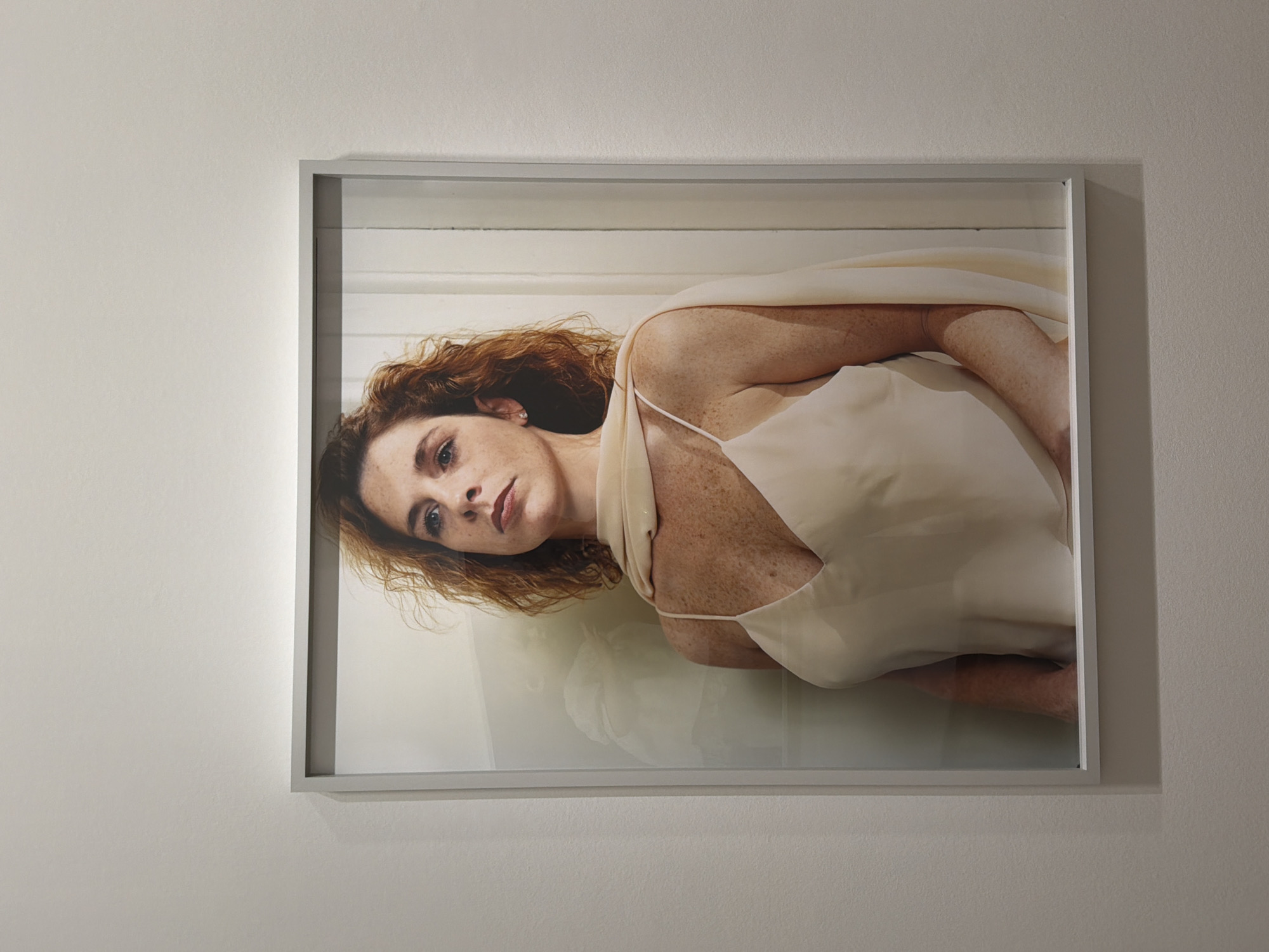A framed portrait of a woman is prominently displayed on a plain, light-colored wall. The large, rectangular photograph, presented in a landscape orientation, is enclosed within a simple, light grey or silver frame. The subject is a woman with reddish-brown, wavy hair, fair skin, and numerous freckles visible across her face, neck, and shoulders. She has blue eyes and a calm, direct, yet somewhat contemplative expression. She is depicted lying down, or in a reclined pose, with her head turned slightly towards the viewer. She wears a light-colored, possibly cream or off-white, sleeveless dress with delicate spaghetti straps, and a sheer, flowing piece of fabric draped over her shoulders. A subtle earring is visible in her left ear. The background within the portrait is a light, muted tone, with a faint reflection appearing beneath her body, suggesting a smooth, possibly reflective surface like water or polished stone. The photograph is well-lit, highlighting the textures of her skin and hair. This display is situated in Paris, France. There is no visible text on the image or the surrounding wall.