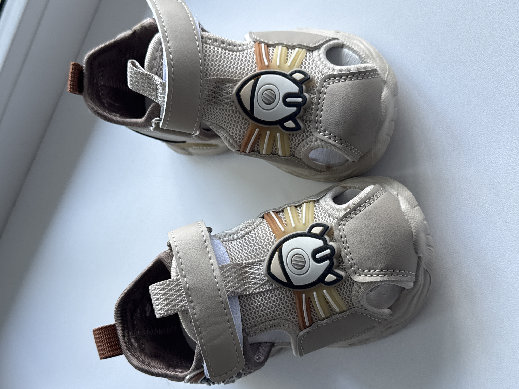 The image shows a pair of beige children's sandals with a rocket-shaped applique on each side. The sandals have a velcro strap closure and are made of mesh and synthetic leather. They are placed on a white surface, likely a windowsill, with a light blue background that suggests an outdoor environment or a bright sky. The sandals appear to be new and in good condition. The context of "Яромінскі сельскі Савет, Belarus" suggests this photo was taken in a rural area within this administrative division of Belarus.