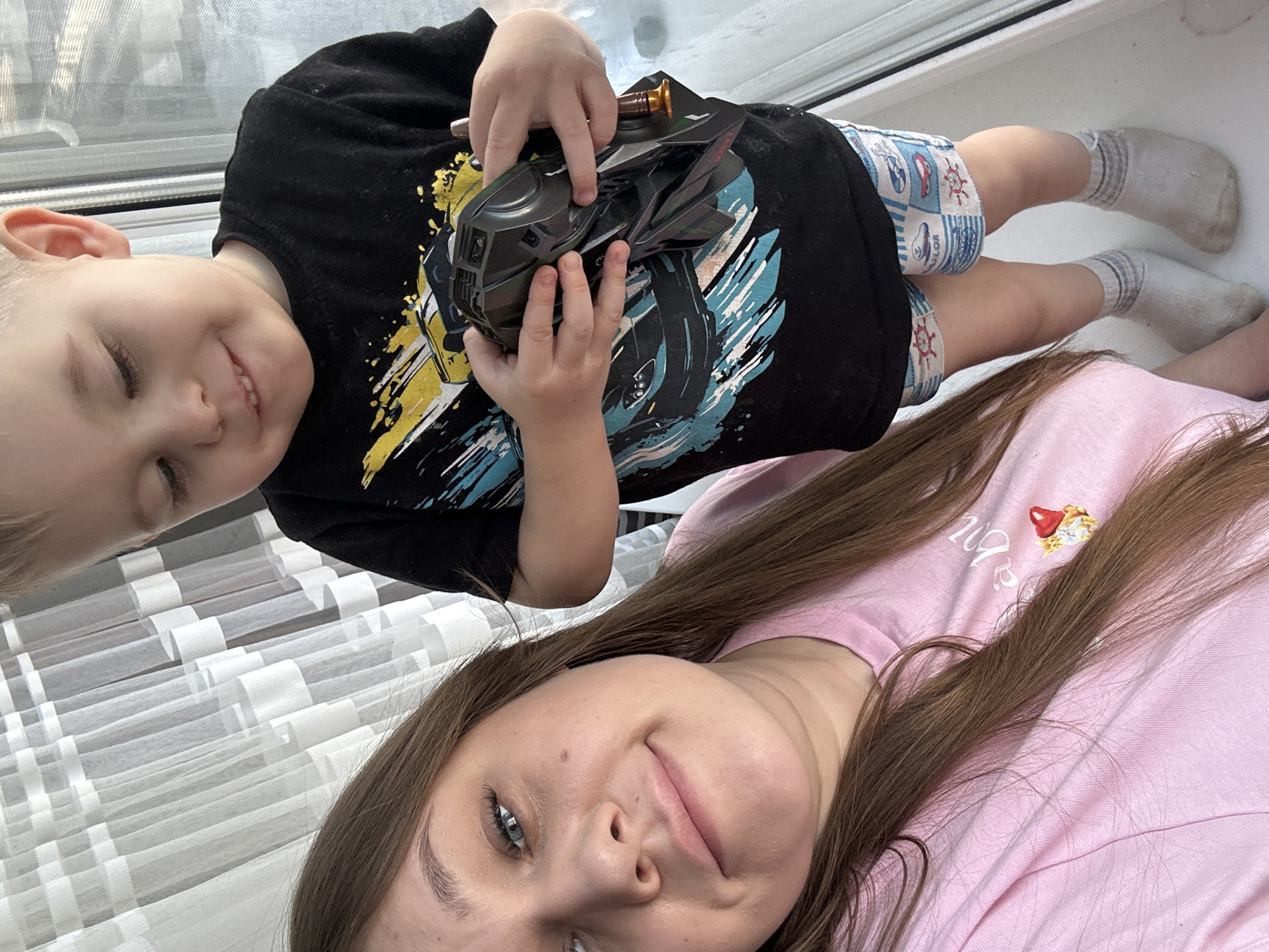 A tilted, close-up selfie-style image captures an adult woman and a young child indoors, likely by a window. The woman, positioned in the foreground and closer to the camera, has long brown hair falling over her shoulders and a light pink t-shirt. She has blue eyes and a faint, pleasant smile, looking directly at the camera. On her shirt's upper left chest, there's a small design featuring a red-capped object (possibly a mushroom or a dessert) with some white, partially illegible text underneath, which might read "baby" or "happy" or similar.
Beside her, a young boy is visible in the upper part of the frame, smiling broadly with his eyes slightly squinted in delight. He has light blonde hair and is wearing a black short-sleeved t-shirt with a vibrant, abstract graphic design in yellow, blue, and white. His shorts are blue and white with a nautical pattern of anchors and small ships. He is wearing white socks with grey stripes around the ankles. The boy is holding a dark, futuristic-looking toy, possibly a vehicle or weapon, with intricate details and angular shapes. His small hands are wrapped around the toy, suggesting he is actively playing with it.
The background shows a window with a light-colored, possibly sheer, curtain featuring a distinctive pattern of horizontal and vertical bands, creating a grid-like or ladder-like effect. Natural light streams in from the window, indicating it is daytime. The overall mood of the image is joyful and relaxed, capturing a personal moment between the two individuals.
