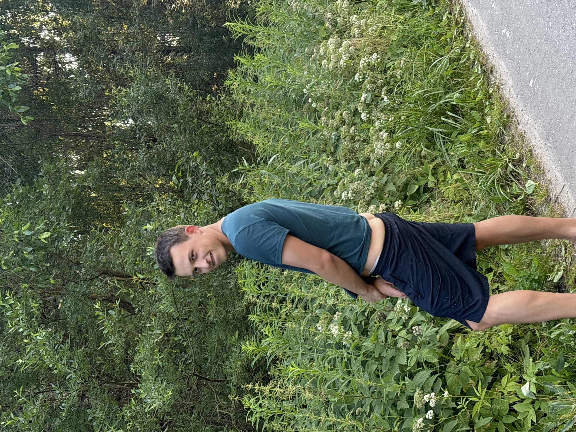 A young man in a teal t-shirt and navy shorts is standing in front of dense green foliage, with a paved path to his right. He is facing away from the camera and looking over his left shoulder with a slight smile. His right hand is obscured by his shorts. The background is dominated by lush green trees and smaller plants with white flowers. The lighting suggests it is daytime, possibly late afternoon.