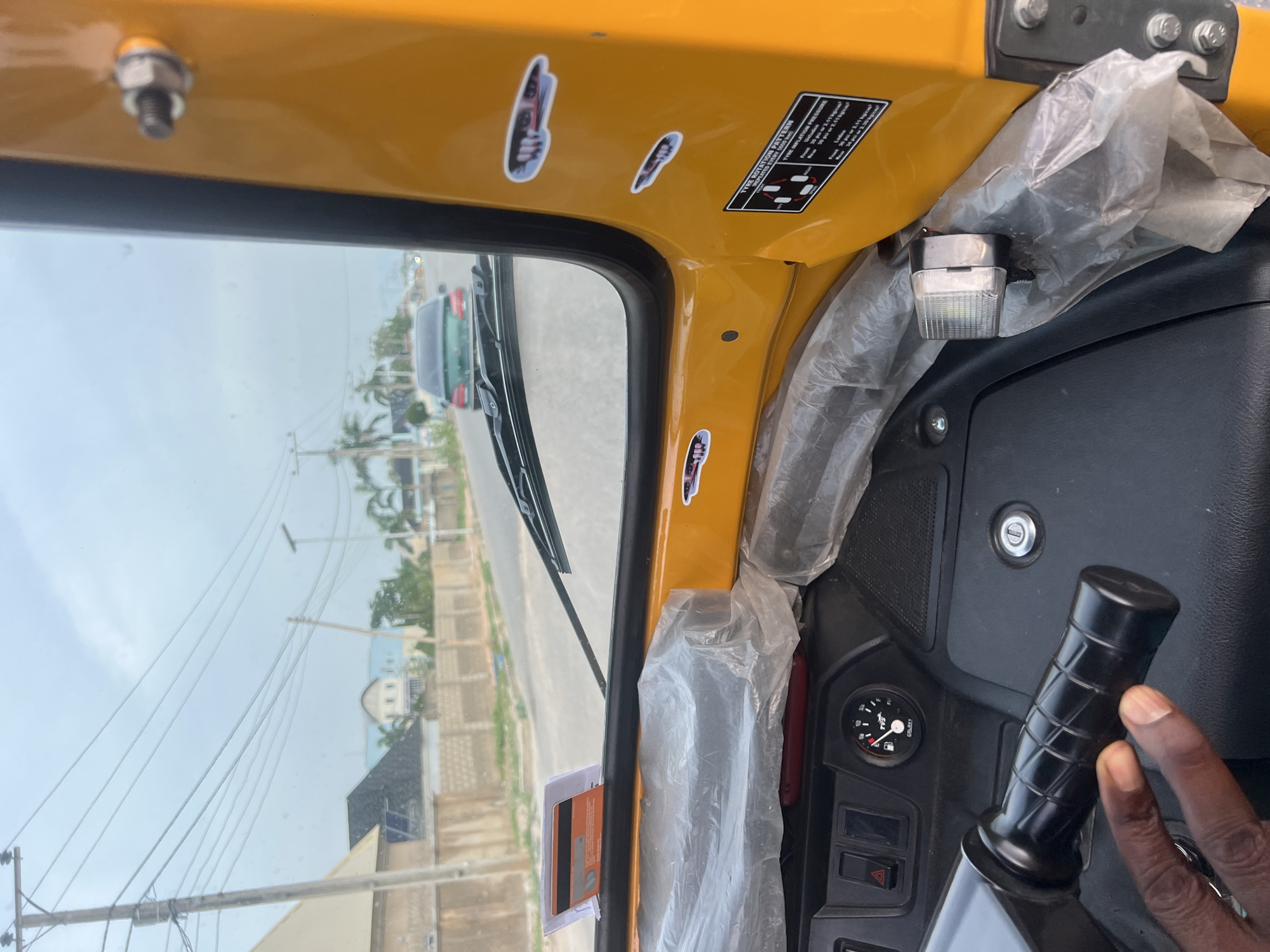 The image captures an interior view from a yellow tricycle, looking out through the front windshield onto a street scene. The perspective is from the driver or passenger seat, with a dark-skinned hand gripping a black ribbed handlebar in the lower right foreground. The vehicle's interior is bright yellow, and parts of the dashboard and a small LED light fixture are covered in clear plastic sheeting.
On the yellow frame above the windshield, a sticker provides "TYRE ROTATION PATTERN" information, indicating "REPEATED EVERY 5000" and "5000km)", along with a diagram. Below this, "TYRE INFLATION PRESSURE," "Laden 34," "02.11," and "Untadien" are visible. Other small, decorative stickers depicting hands holding what appear to be knives are also present. A small dashboard gauge is partially visible.
Through the windshield, a paved road can be seen, bordered by utility poles with power lines and scattered palm trees. Various buildings, including one with a prominent white dome, are in the background. Another dark-colored vehicle is visible further down the road. The sky is overcast, indicating daytime. The scene depicts typical urban or semi-urban transportation in Howayi, Nigeria.