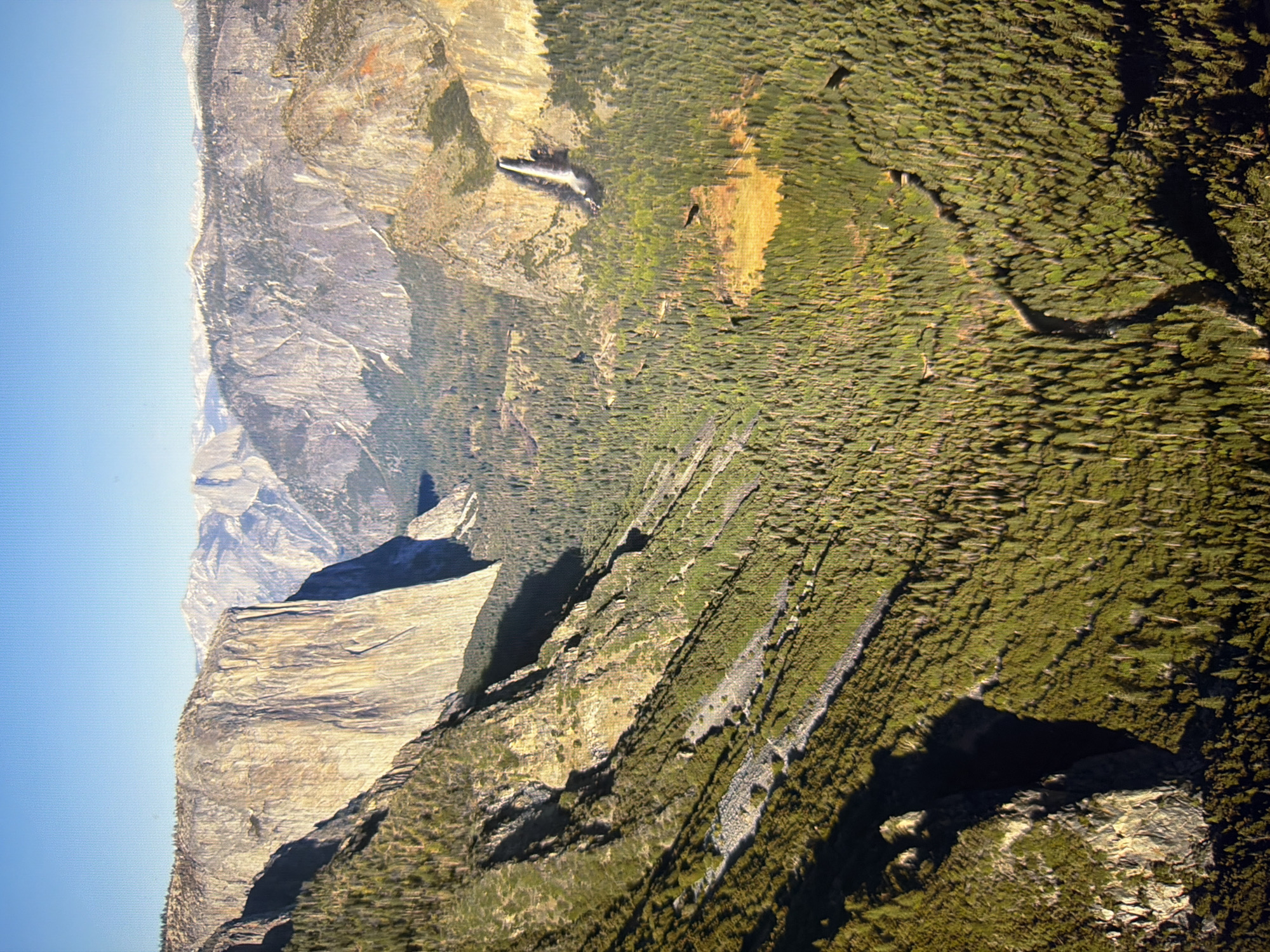 This is an aerial, wide-angle photograph of Yosemite Valley. Towering granite cliffs dominate the left and upper portions of the image, with El Capitan being a prominent feature on the left. The valley floor is covered in dense coniferous forest. A waterfall, likely Yosemite Falls, is visible in the distance on the right, cascading down a cliff face. The sky is clear and blue. The image does not depict any part of the city of Kyiv, Ukraine.