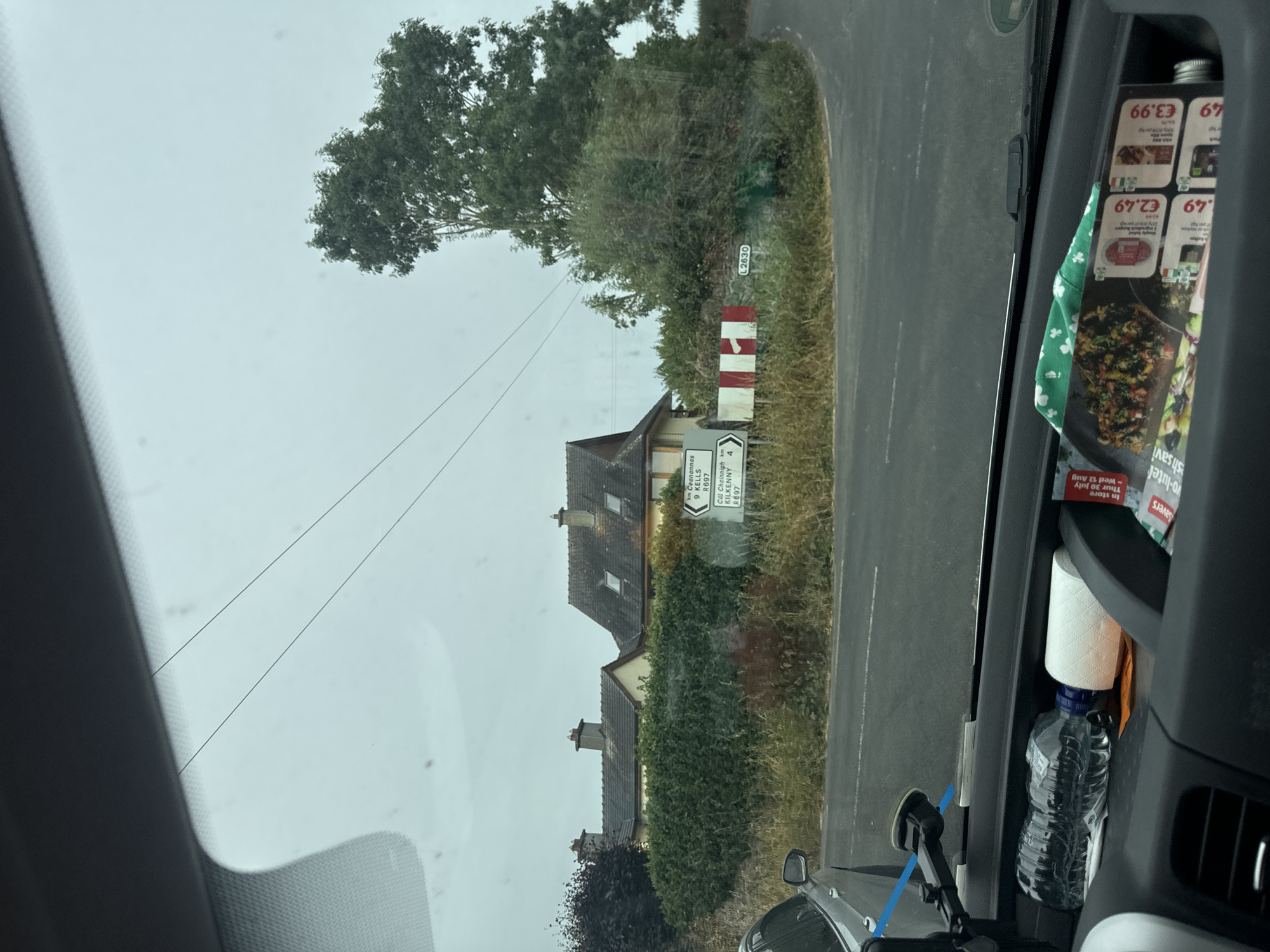 The image shows a view from inside a vehicle looking out at a rural intersection. The intersection features a white signpost with directional arrows and text indicating "km Ceanannas 9 KELLS R697" and "Cill Cheannain R697". There are also smaller signs indicating "L2630" and a red and white striped marker. In the background, two houses with tiled roofs are visible behind a dense green hedge. A mature tree with dark green foliage stands to the right of the intersection. The sky is overcast and appears light gray. The foreground shows the interior of the vehicle, including a water bottle, a roll of toilet paper, and some promotional flyers or magazines with prices displayed in Euros. A silver car is partially visible on the left side of the frame. The road surface is asphalt.
