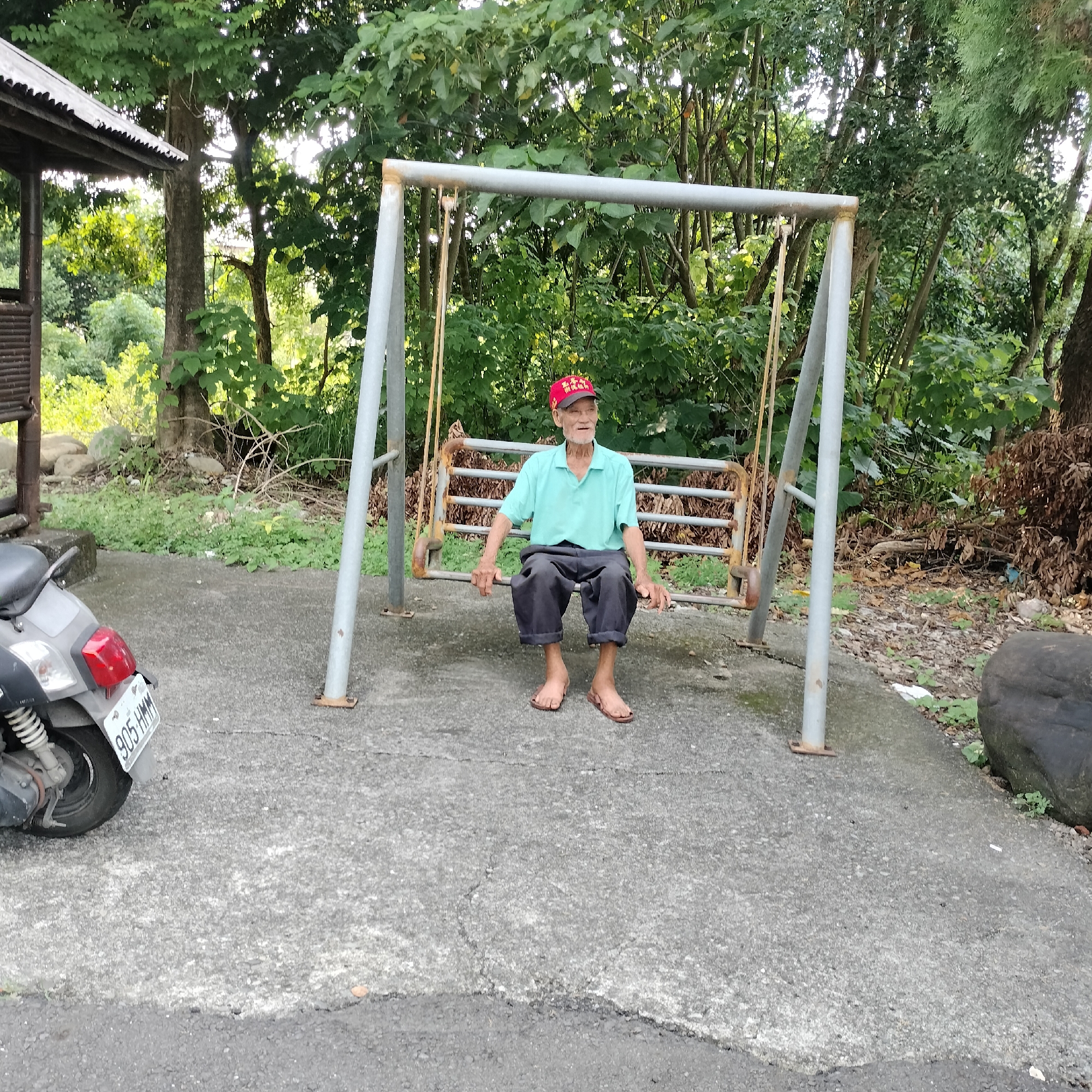 An elderly man wearing a light blue polo shirt, dark pants, and a red cap is sitting on a rusted swing set in an outdoor setting. The swing set is constructed with gray metal poles and a metal bench. The man is facing forward and looking to his right with a slight smile. Behind the swing set, there is dense green foliage and trees, suggesting a natural or park-like environment. To the left of the swing, a portion of a simple outdoor structure with a corrugated metal roof is visible, and a gray scooter with a license plate reading "905-HMM" is parked on a concrete ground. The ground is a mix of concrete and dirt, with some scattered leaves and small rocks. The overall scene appears to be in a rural or semi-rural area, consistent with Zhushan Township, Taiwan, given the context.