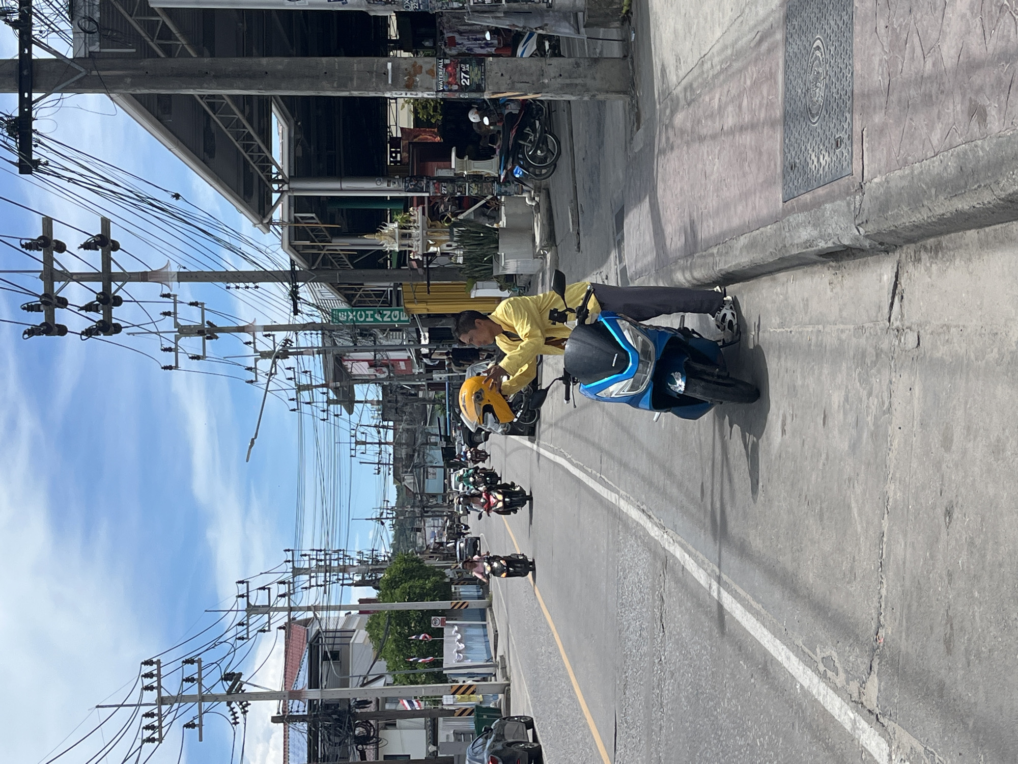 The image depicts a street scene in Tambon Ko Pha Ngan, Thailand, under a partly cloudy sky. A man in a yellow shirt and gray pants stands next to a blue and black scooter, holding a yellow helmet. He appears to be interacting with the helmet, possibly preparing to wear it. The scooter is parked on the right side of the road, on a paved sidewalk.
The street itself is asphalt, marked with a double yellow line in the center. Several other scooters and motorcycles are visible on the road, some with riders. In the background, there are buildings, some with awnings and signs, and numerous utility poles with a complex network of wires. The architecture suggests a commercial or residential area. The environment appears to be a typical street in a Thai town, with evidence of bustling activity.