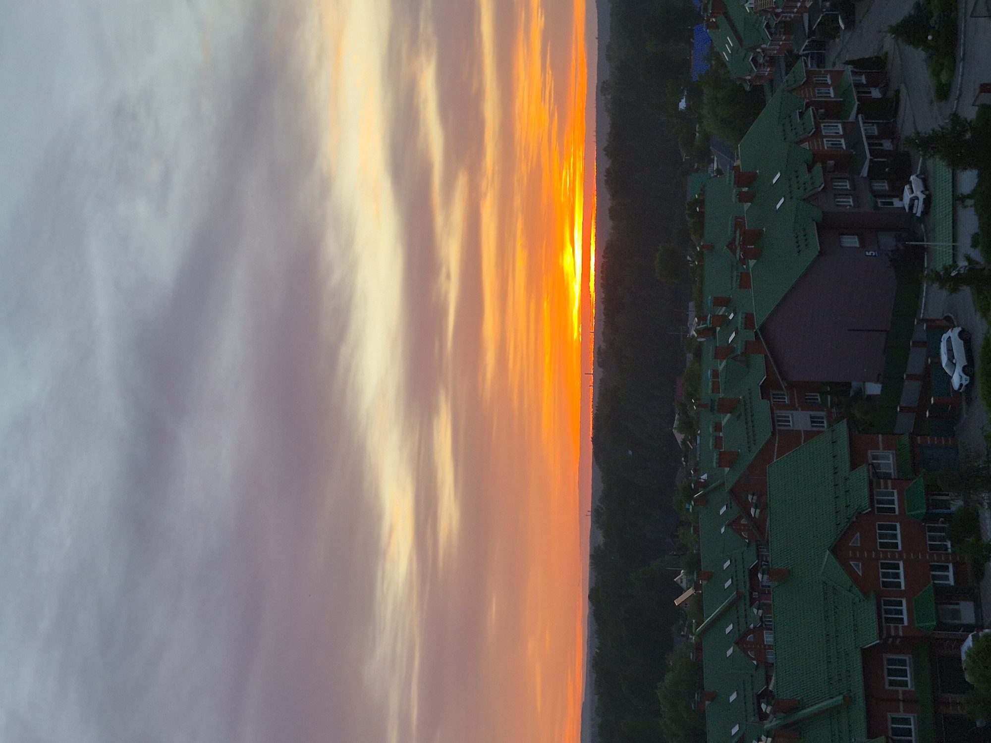 A view of an unknown city at sunset, viewed from a high angle. The sky shows an array of colors, from shades of pale purple to orange and golden hues near the horizon where the sun appears to set. Below the horizon is a dense line of trees. On the right side of the frame, residential buildings with green rooftops can be seen. There are also some white cars parked beside the houses.