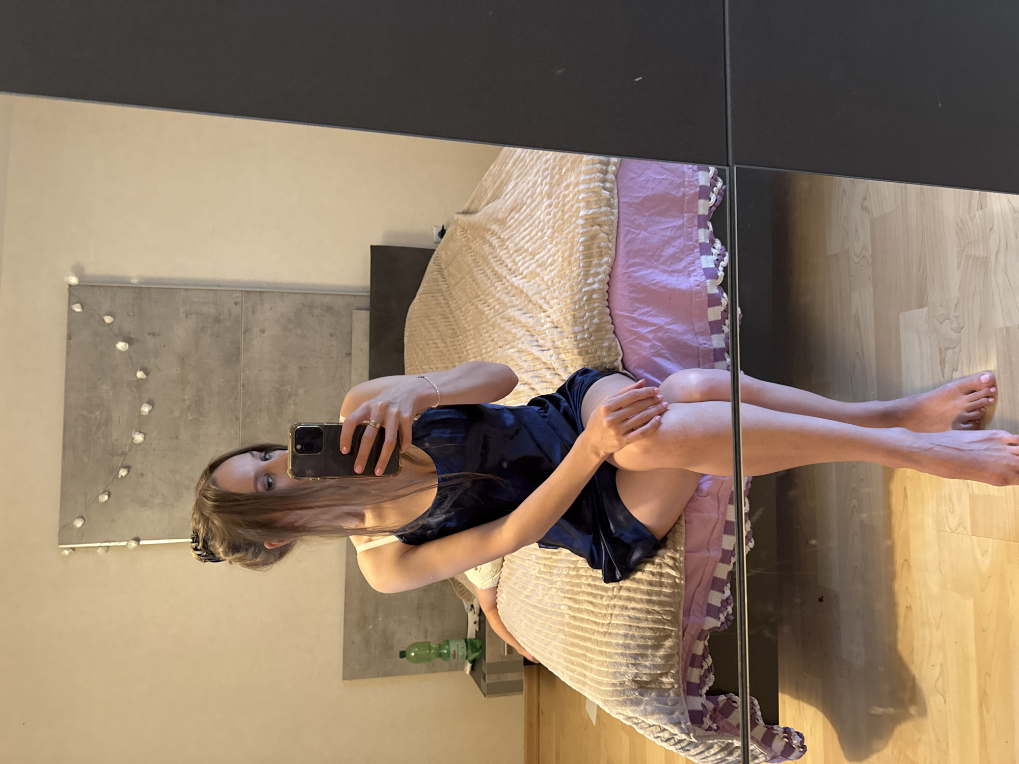 A young Caucasian woman with long brown hair styled in a bun, wearing a dark blue slip dress, is sitting on the edge of a bed in a bedroom. She is holding a smartphone in her right hand, taking a mirror selfie. Her left arm is resting on her lap. The bed is covered with a textured beige blanket and a pink sheet with a white and purple trim. To the left of the bed, on a nightstand, is a green plastic bottle. The background shows a textured wall with decorative lights. The floor is made of light-colored wood. The scene appears to be an interior residential setting, likely a bedroom. No specific indicators of the city of Überherrn, Germany are visible in the image.