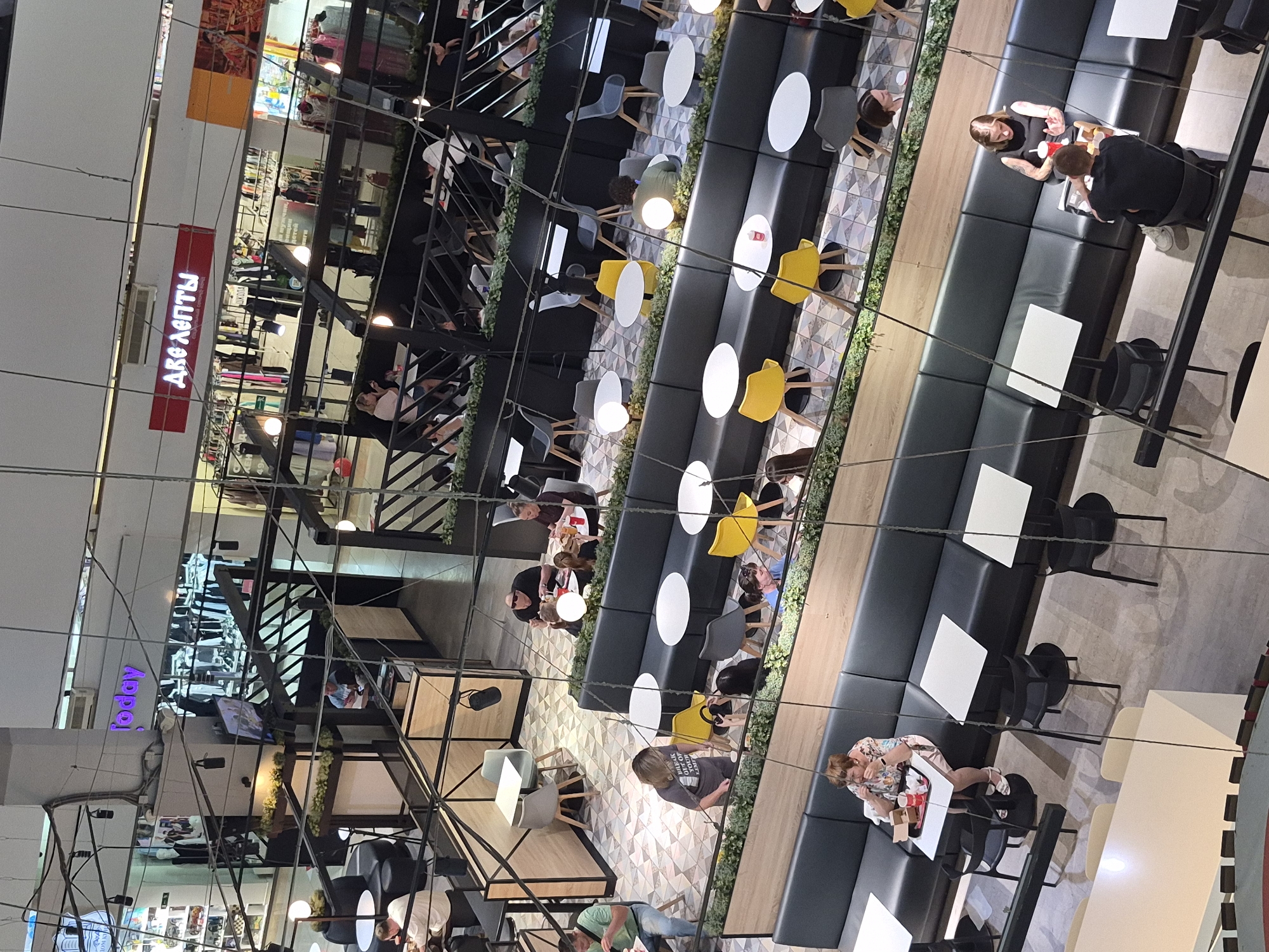 The image shows an interior view of a multi-level shopping mall, likely in Yekaterinburg, Russia, given the visible Cyrillic signage. The scene is captured from an elevated vantage point, looking down onto various dining areas and retail spaces.
In the foreground, a seating area with black leather banquettes and white, round tables is occupied by several individuals. One woman, wearing a floral dress, is seated at a table with a food tray. Another table is occupied by a woman with tattoos on her arms and a man facing away from the camera.
Further back, other seating areas are visible, with a mix of yellow and grey chairs around white tables. Some individuals are seated and appear to be dining or conversing.
Above the dining areas, retail spaces are visible. One sign prominently displays Cyrillic text reading "две лепты" (two kopecks). Another sign in purple reads "today". The retail areas feature clothing racks and mannequins.
The overall environment appears to be a modern shopping mall with various amenities, including a food court or restaurant section. The lighting is a mix of ambient mall lighting and artificial light from fixtures within the dining areas and shops. The floor in the dining area has a geometric tile pattern. Overhead structures with black metal framing and wiring are also prominent in the composition.