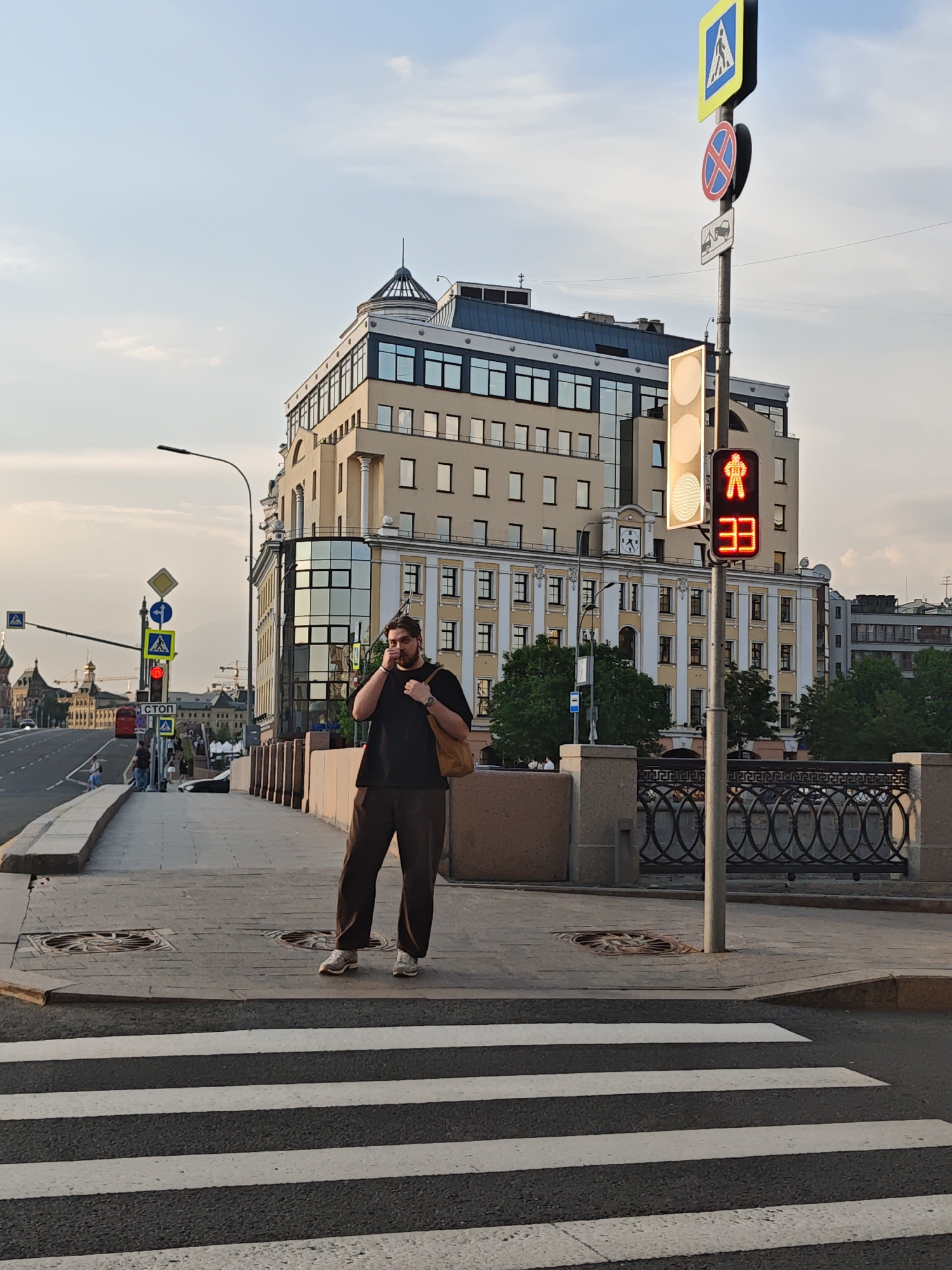 A person is standing on a crosswalk in an urban setting in Unknown, Unknown. The person is wearing a black shirt and brown pants and is holding a cell phone to their ear. They also have a brown bag slung over their shoulder. In the background, there is a large building with a beige facade. A traffic light shows a red walking man symbol with the number 33. Various road signs are also present. The sky is light blue with some clouds.