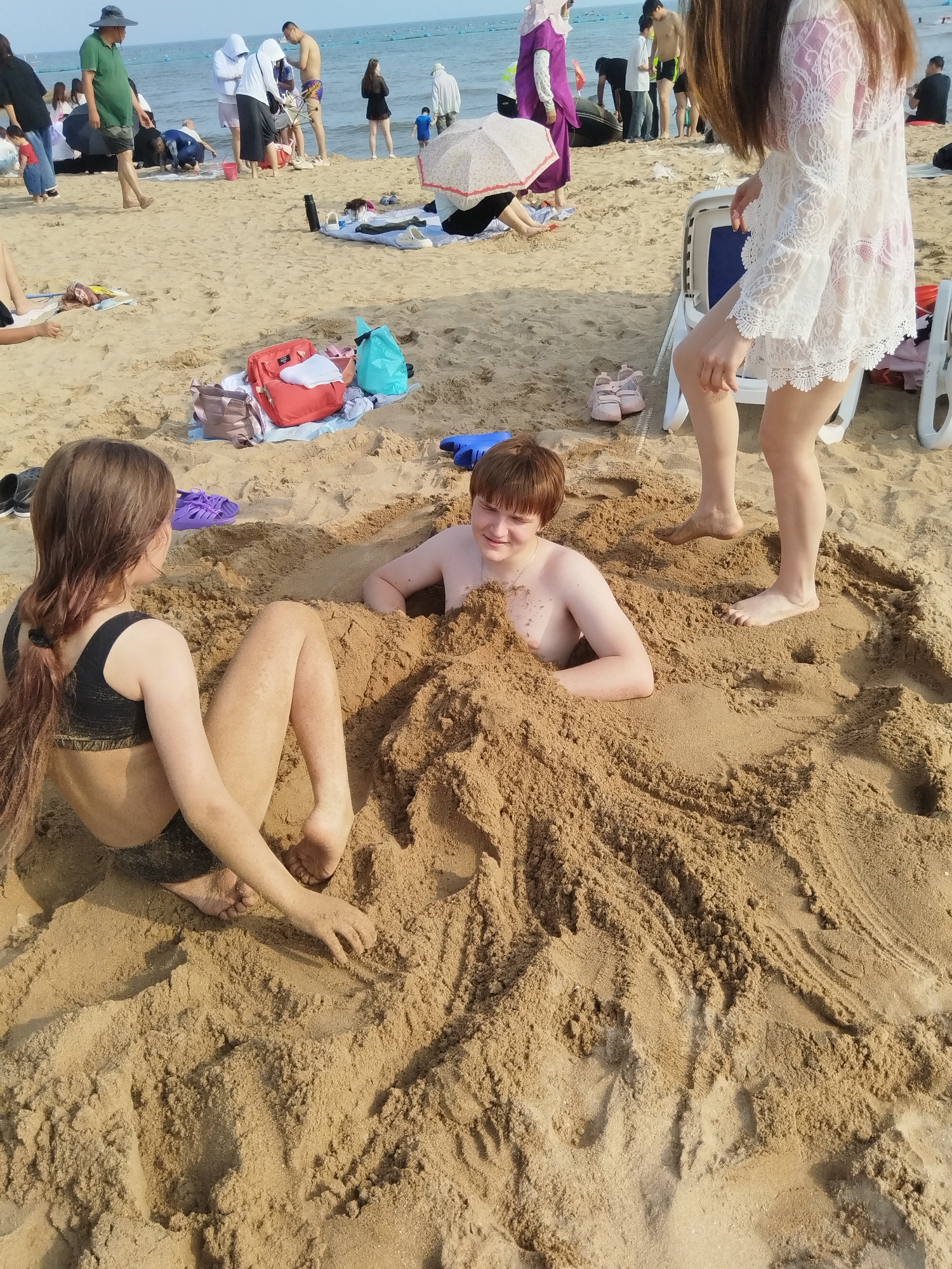 The image depicts a sunny day at a beach in Qinhuangdao, China, with the ocean visible in the background. The focus is on two children partially buried in the sand. On the left, a girl with long brown hair, wearing a black bikini top and bottoms, is sitting with her legs extended. On the right, a boy with short brown hair and a necklace is lying down, submerged in the sand up to his chest. Other beachgoers and various objects like bags, shoes, and beach umbrellas are scattered around the scene. The overall atmosphere appears relaxed and playful, suggesting a family outing or a day of recreation at the beach. The time of day is likely midday or early afternoon, given the bright sunlight and the shadows cast on the sand.
