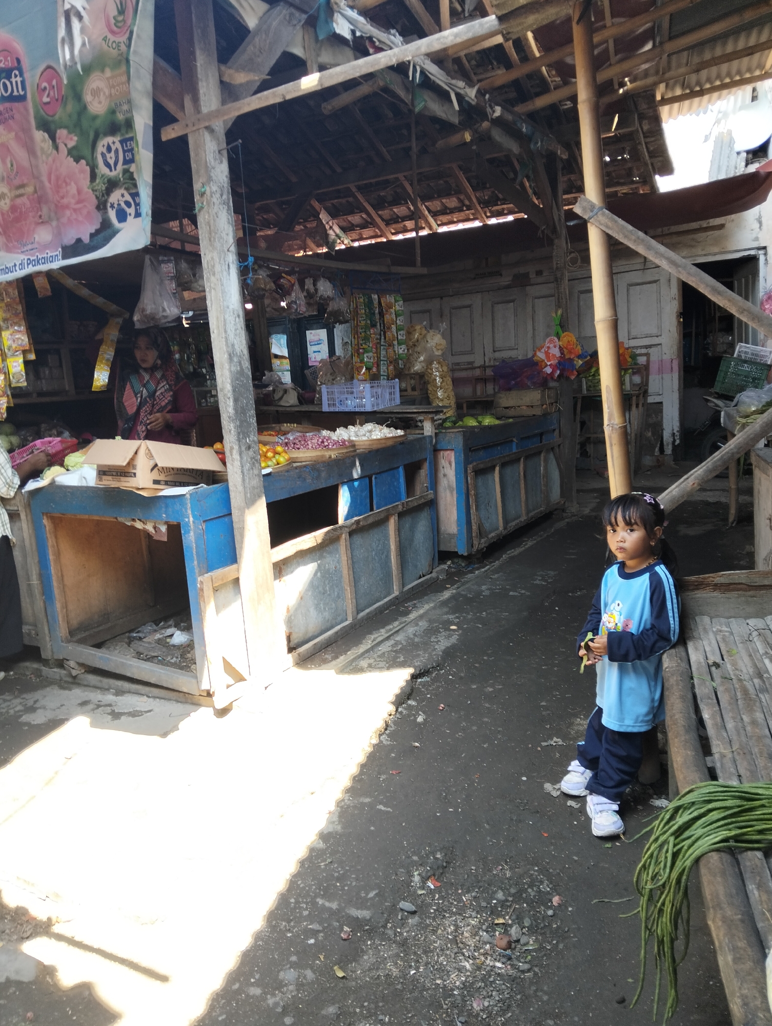 A young girl with dark hair, wearing a light blue long-sleeved shirt and dark blue pants, stands next to a wooden bench, holding a bunch of green beans. She is barefoot and appears to be looking at the camera. The scene takes place in an outdoor market in Muncar, Indonesia, likely a fish market given the location's prominence. In the background, a covered stall displays various goods, including produce like onions and tomatoes, packaged snacks, and cleaning products. A woman in a patterned headscarf and maroon top is visible behind the stall. The market structure is rustic, with wooden beams and a corrugated metal roof. The ground is a mixture of dirt and pavement, with some debris scattered around. Sunlight casts strong shadows across the scene, indicating it is daytime. The overall impression is of a bustling, open-air market with a mix of goods and people.