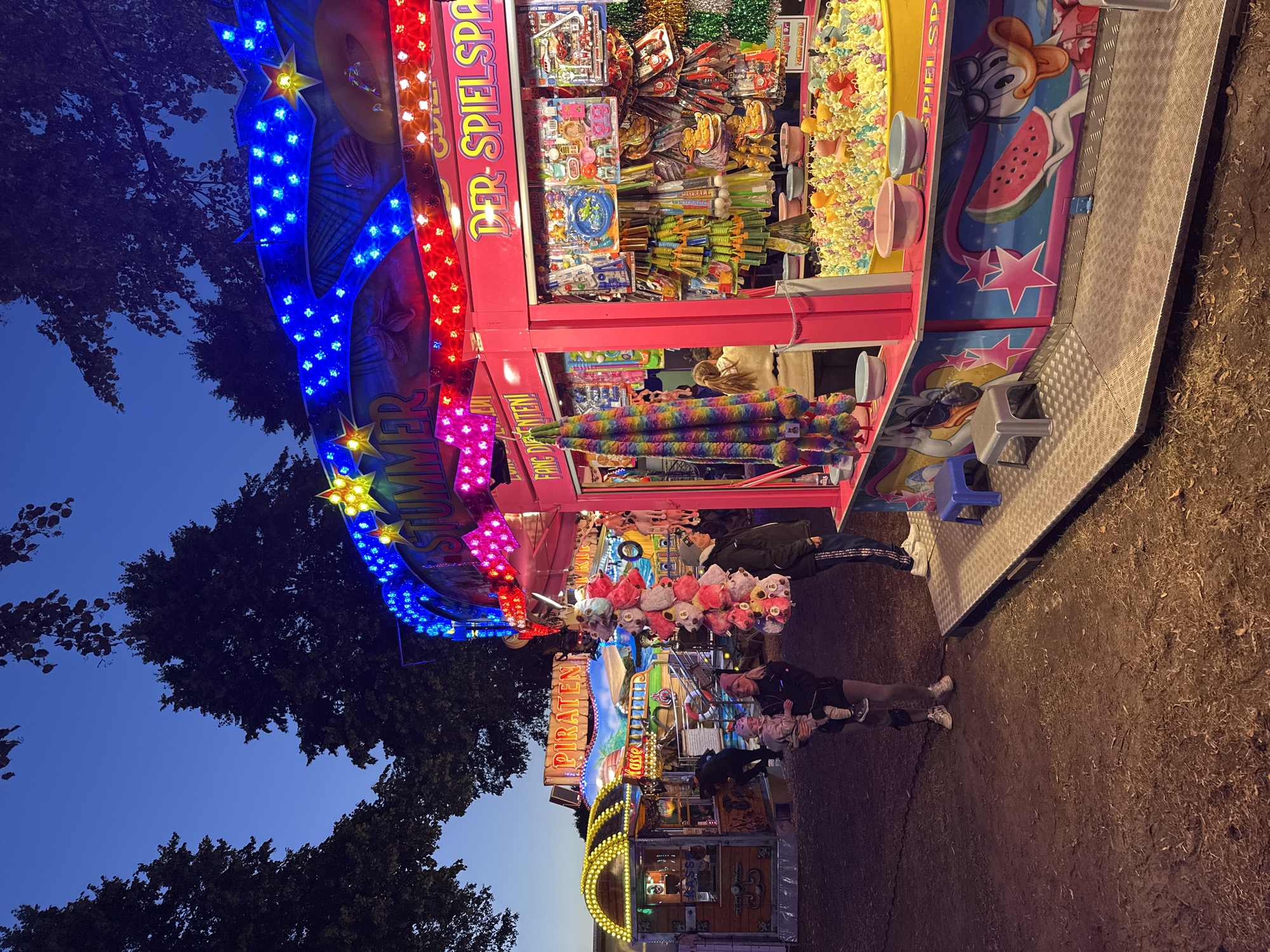 The image depicts a scene at a fairground in Bruchhausen-Vilsen, Germany, at dusk. Several amusement stalls are visible, illuminated by colorful lights. In the foreground, a stall brightly lit with pink and blue LEDs features prizes such as plush toys hanging from the front, and a game involving rubber ducks in a pool. A woman holds a baby, standing near this stall, while another person stands to her right. To the left, another stall with yellow lights has a sign that reads "PIRATEN" (Pirates) and "KASSE" (Cashier). The ground is dirt and scattered with leaves, indicating an outdoor setting. The overall atmosphere suggests a lively evening event.