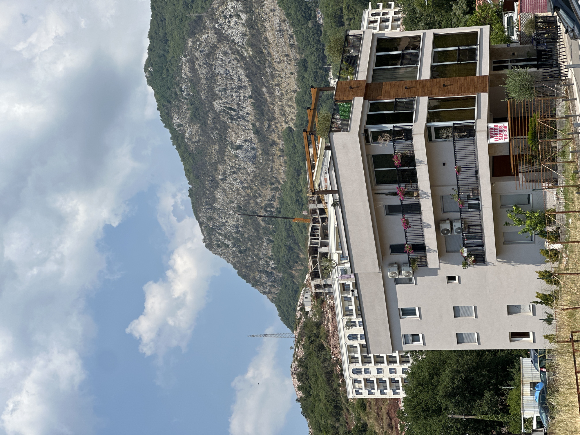 This media file captures a view of a modern, multi-story building situated on a hillside, with a steep, rocky mountain covered in dense green vegetation dominating the background. The sky is partly cloudy, with bright white clouds scattered across a pale blue expanse, suggesting it is daytime with fair weather. The building exhibits a contemporary architectural style, featuring large windows, balconies with black railings, and light-colored facade. Decorative wooden accents are present on the upper floors. Air conditioning units are visible mounted on the exterior walls. A red and white sign is attached to the building, displaying text that appears to be in a Cyrillic alphabet. In the distance, a large construction crane is visible, indicating ongoing development or renovation in the area. To the right of the building, a wire fence runs along the property line, and some greenery is present. The scene is set in a mountainous region, likely a residential or commercial area, given the proximity of the building to the natural landscape.