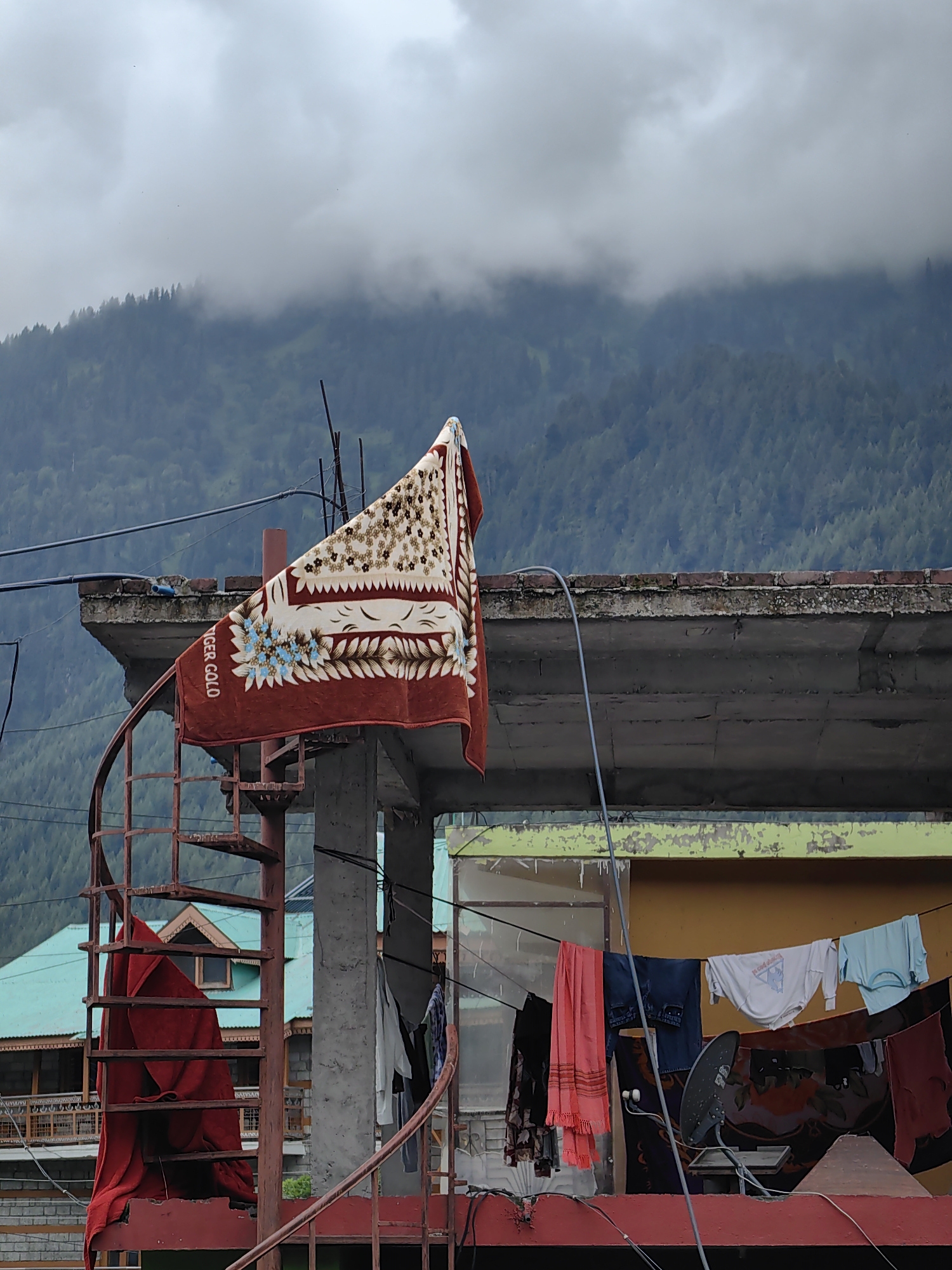 The image depicts a rooftop scene in Barheta, India, under a cloudy sky with a misty mountain range in the background. A spiral staircase, appearing rusted, is partially visible on the left, with a red blanket draped over it. A blanket with a floral pattern, possibly a 'Tiger Gold' blanket, is hung from a pole on a concrete structure. In the foreground, laundry hangs from a clothesline, including a pink scarf, jeans, a white t-shirt, and a light blue t-shirt, along with other dark-colored fabrics. A satellite dish is mounted on the wall of a building behind the clothesline. The architecture suggests a residential area, with buildings made of concrete and brick. The overall atmosphere is overcast and slightly damp, consistent with mountain weather. No people are present in the frame.