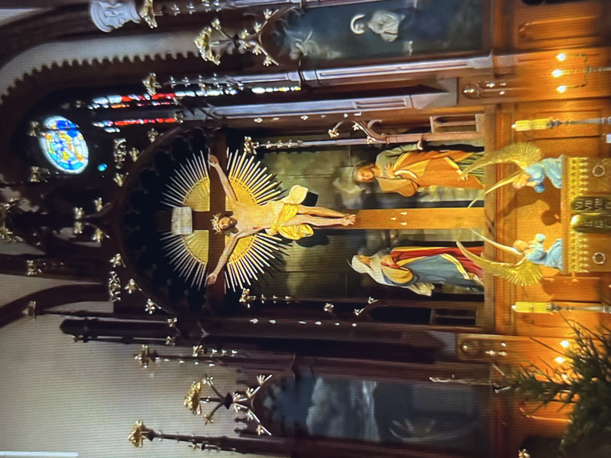 The image depicts the interior of a Catholic church, likely located in Tinaquillo, Venezuela, as per the user’s context. The scene is focused on the altar area, dominated by a large crucifix featuring Jesus Christ with golden rays emanating from behind. To the right of the crucifix, there are statues of figures, possibly representing Mary and John, with angel sculptures interspersed. A stained glass window is visible on the left. The overall decor is ornate, with dark wood and gilded details. A painting hangs above the crucifix.