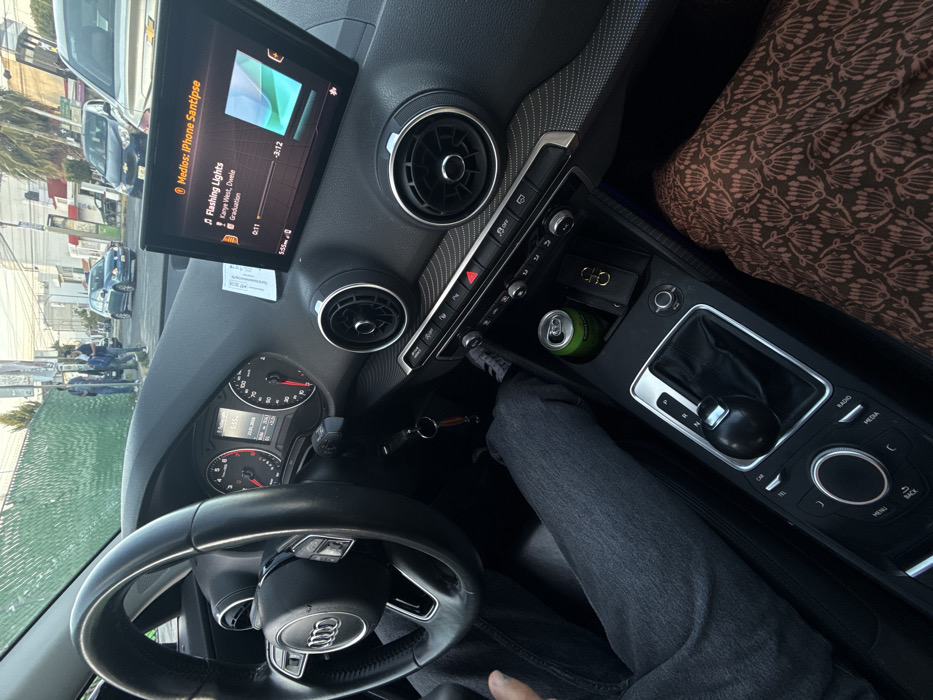 This is an interior shot of an Audi car. The infotainment screen displays the song "Flashing Lights" by Kanye West. The steering wheel has the Audi logo. The center console shows the gear shifter, a can of Sprite, and a "CHO" branded item, possibly a wallet. The person sitting in the driver's seat is wearing grey jeans. Outside, cars and people can be seen through the windshield.