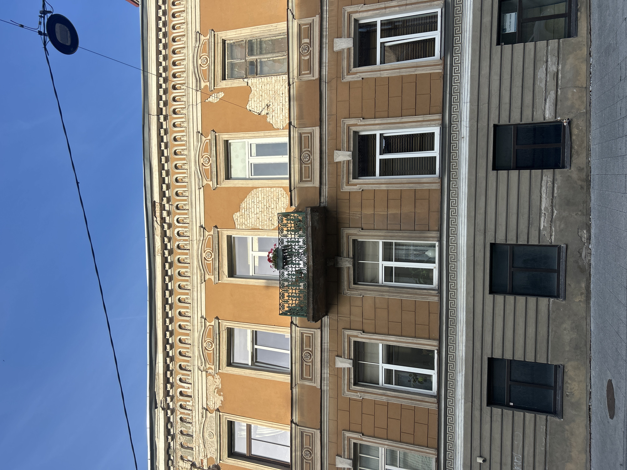 The image captures the facade of an old, multi-story building in Vilnius, Lithuania, under a clear blue sky. The building's exterior exhibits signs of age and wear, with peeling orange plaster revealing brickwork in several areas, particularly on the upper floors. The architecture features classical decorative elements around the windows, including ornate moldings and pediments.
The upper floor has several large windows, some with white frames and blinds, and one with a wrought-iron balcony adorned with a planter of red flowers. The second floor displays a row of identical windows with white frames and blinds. The lowest visible floor is a darker, more muted color with simpler, darker-framed windows. A decorative Greek key border separates the second and lowest visible floors. The sidewalk in front of the building is paved with grey tiles, and a manhole cover is visible. A lamppost with a dark fixture hangs from wires overhead. The overall impression is of an aged urban structure in need of restoration.