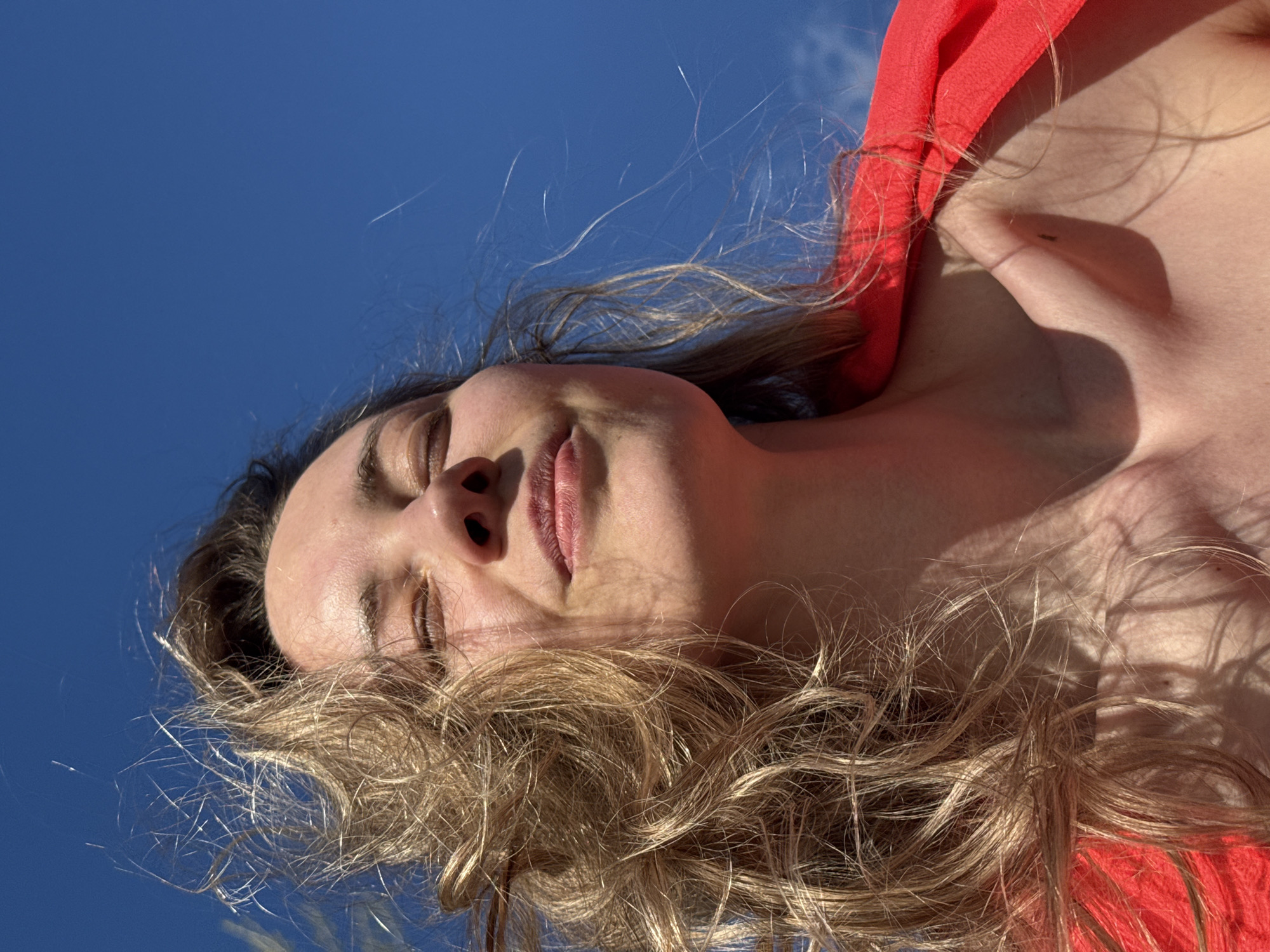 A low-angle shot of a woman, possibly in the city of Chaikovsky, Russia, as requested. She is looking slightly upwards, with her eyes partially closed. The background is a clear blue sky with sparse clouds. She is wearing a red garment, and her blonde hair is spread out around her face, catching the sunlight. The shot appears to be taken outdoors in bright daylight.