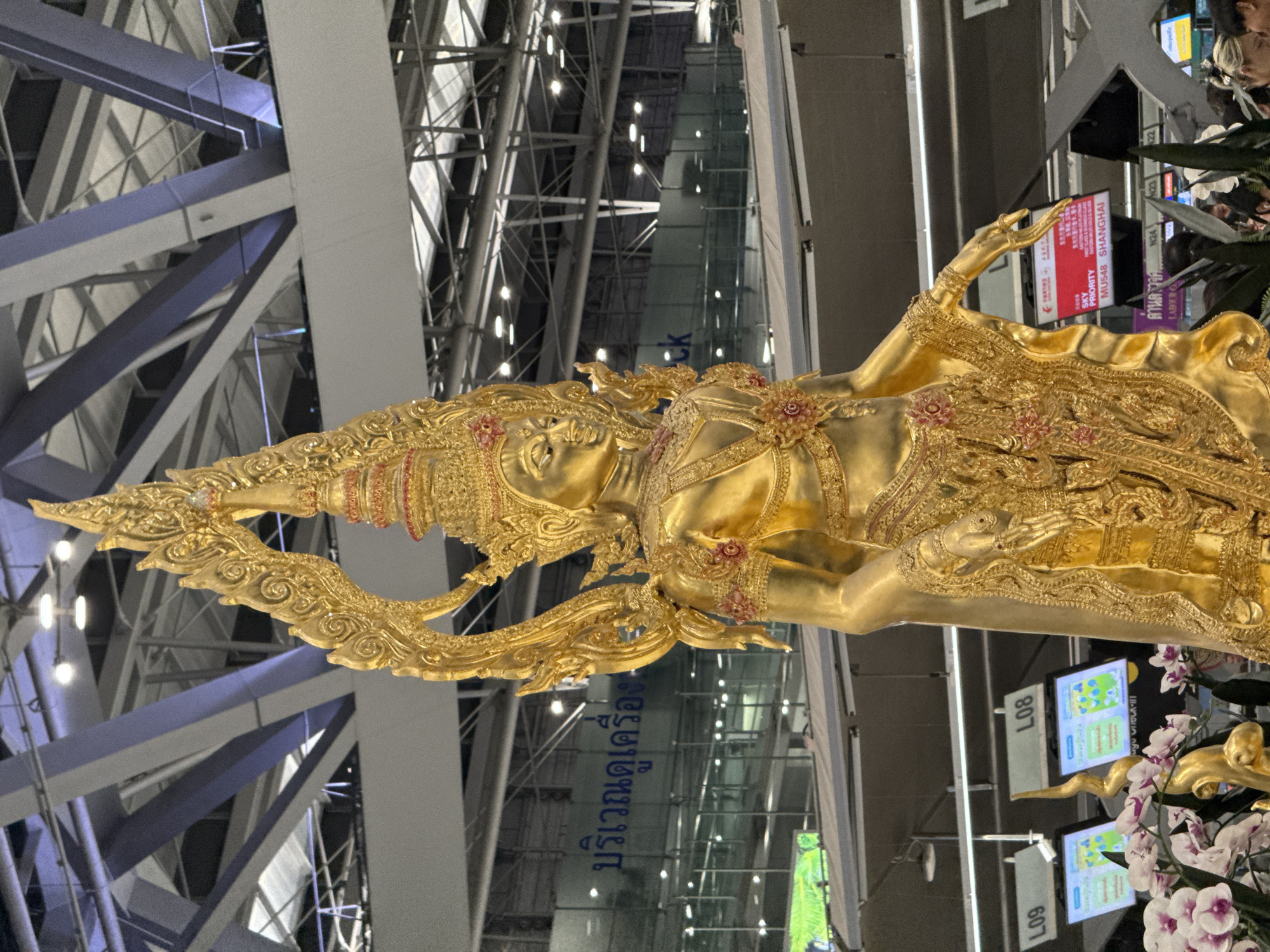 The image shows a large, ornate golden statue of a figure with elaborate headwear and clothing. The statue is positioned in what appears to be an indoor public space, possibly an airport terminal, given the signs and structural elements visible in the background. Signs in Thai script are visible on a building facade behind the statue. In the foreground, there are some orchids and green plants. In the midground, there are several check-in counters with digital displays labeled "L08" and "L09". A red electronic display board in the background shows flight information, including "SKY PRIORITY" and "MU548 SHANGHAI". The overall environment suggests a busy transportation hub. The statue itself is highly detailed, with intricate carvings and decorative elements. The lighting in the scene is artificial, likely from overhead lights.