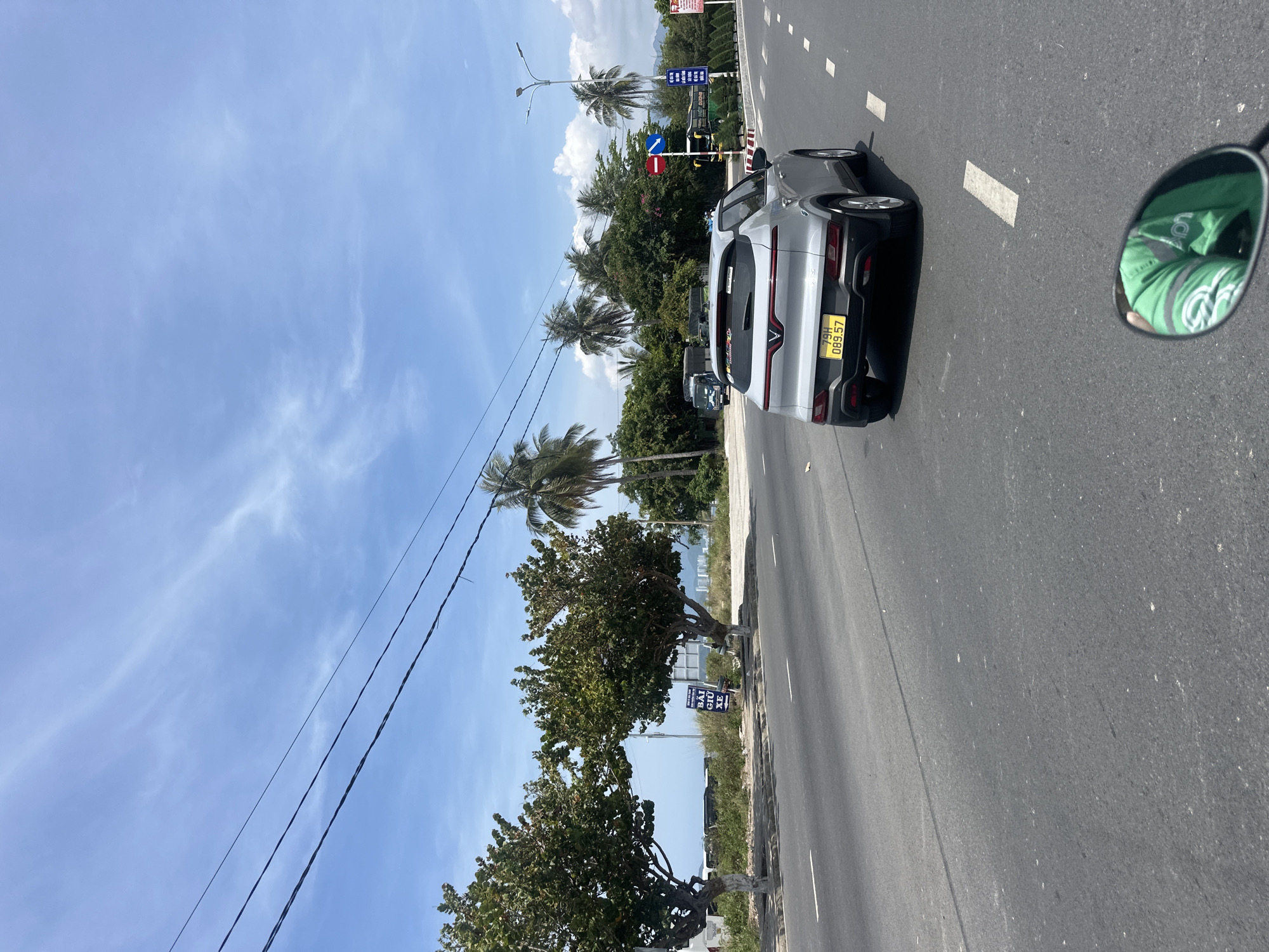 In Bac Nha Trang Ward, Vietnam, a silver VinFast electric car, license plate 79H-089.57, is seen from behind on an asphalt road. The car is in motion, with its brake lights on. The road has white lane markings. To the left of the road, there are trees and a sidewalk. In the background, there are more trees, including palm trees, and buildings in the distance. The sky is blue with some clouds. A motorcycle's rearview mirror is visible in the foreground, reflecting a person wearing a green jacket. Traffic signs indicating a red circle with a diagonal bar and a blue arrow are visible on the right side of the road.