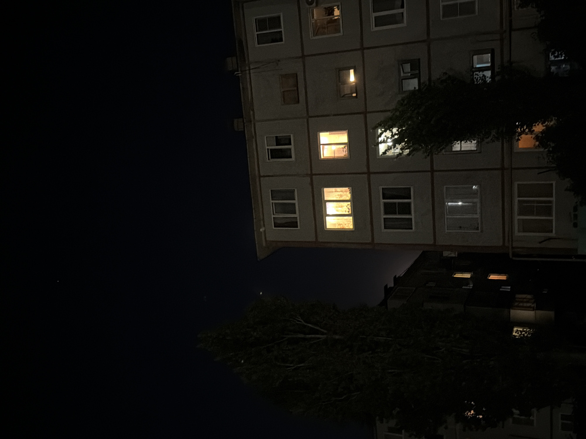 The image shows an exterior nighttime view of apartment buildings in the city of Kamyshin, Russia. The sky is dark, with a few faint stars visible. To the left, a tall, dark tree stands in silhouette against the sky and a slightly darker apartment building. The building on the right is a multi-story apartment block with numerous windows. Several windows are illuminated from within, casting a warm glow. One window on the middle floor of the right building shows a curtain with a patterned design. Another window, to the right of the illuminated ones, appears to be dark. The building's facade is a neutral color, likely concrete or stucco. The overall impression is a quiet, urban night scene.