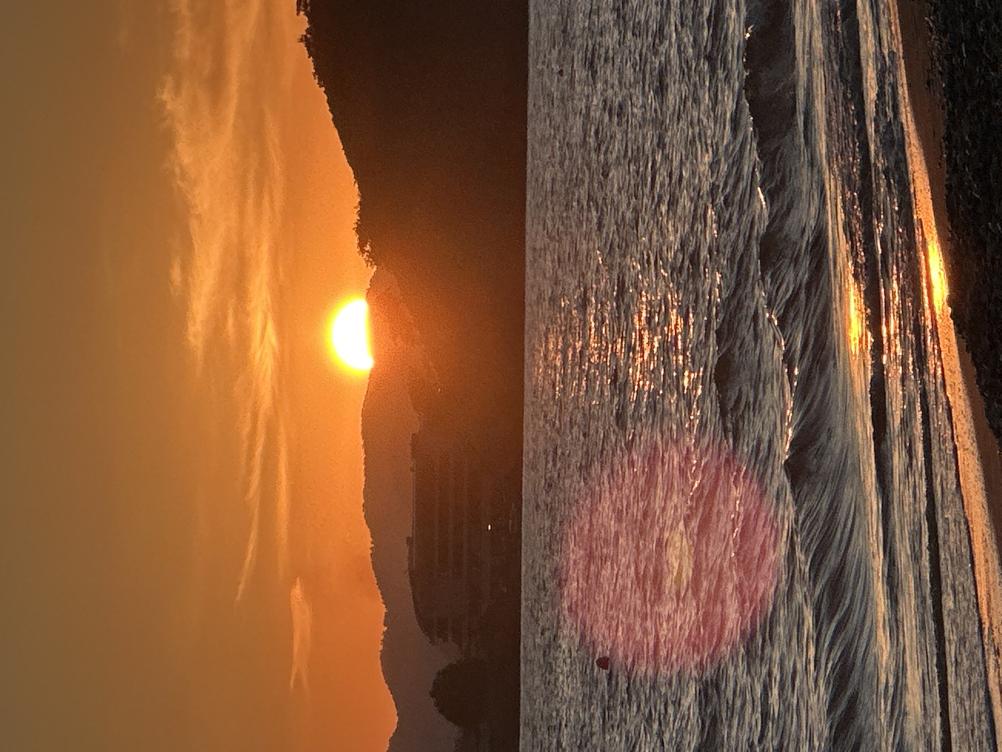 The image shows a sunset scene split into two sections. The left section depicts the sun partially obscured by what appears to be a landmass, casting a golden glow across the sky. The right section shows water with reflections of the setting sun, indicating a coastal area. The reddish hue suggests the lighting conditions associated with sunset. The location is likely Fethiye, Turkey, as specified, though specific landmarks are not visible. The scene consists of natural elements – sun, sky, land, and water. There are no people visible in the image.