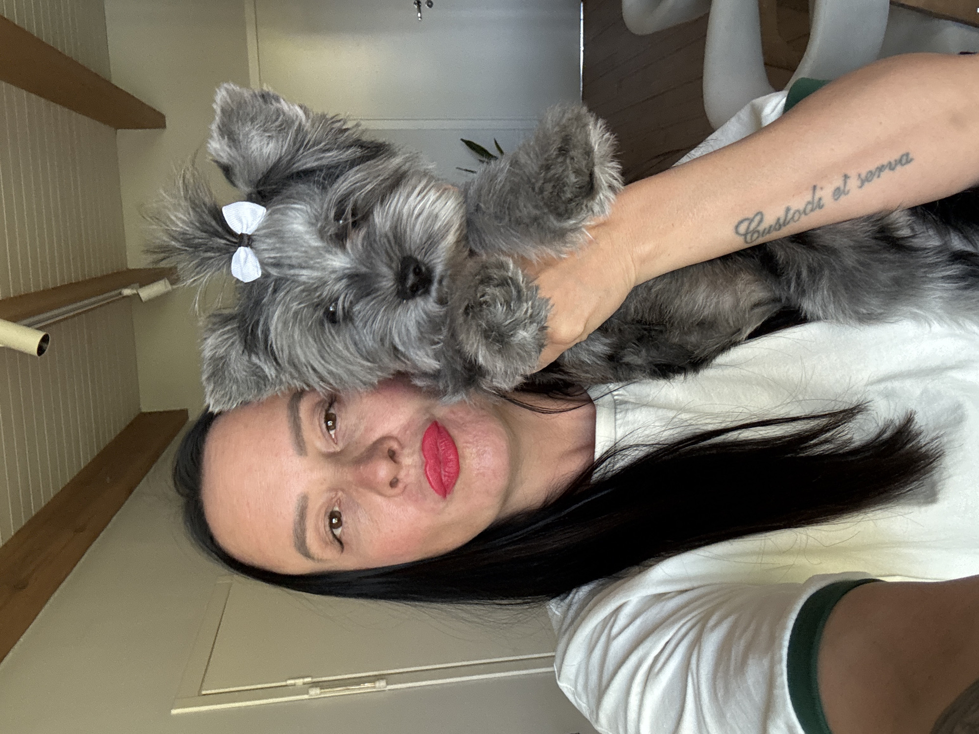 A selfie depicts a woman holding a grey Schnauzer dog. The woman has dark hair, fair skin, and is wearing red lipstick. The dog has a white bow in its hair. The woman is wearing a white t-shirt with a green trim. The background includes a white door, a wooden beam, and a white chair. The location is inferred to be Chaikovsky, Russia.