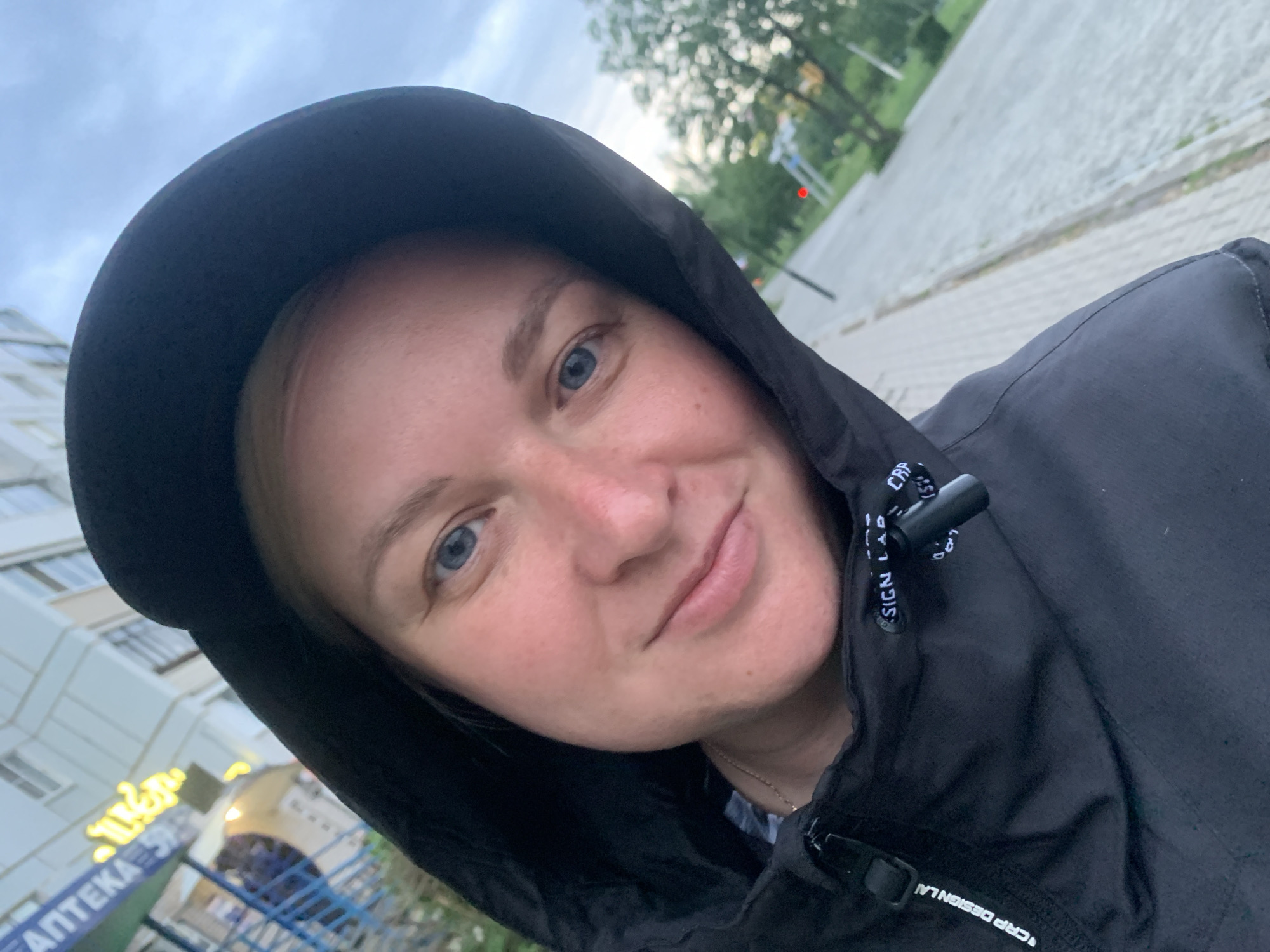 A close-up photograph taken in Chaikovsky, Russia, features a person with fair skin and blue eyes, wearing a black hooded jacket. The hood is up, partially obscuring the hairline. The person is looking directly at the camera with a slight smile.
In the background, on the left, is a building with a sign in Russian that includes the word "АПТЕКА" (pharmacy). The foreground also shows blue metal bars, possibly a fence. The background indicates an urban setting with a sidewalk and greenery. A traffic light post is visible further in the background.