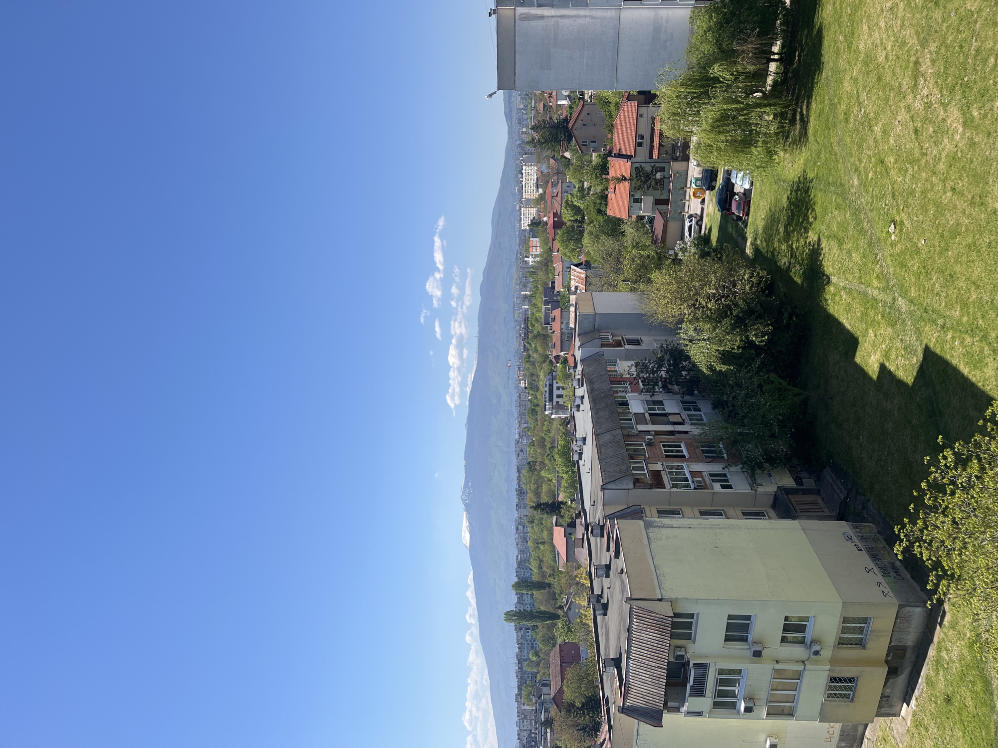 The image shows an elevated view in Sofia, Bulgaria, featuring urban buildings, vegetation, and distant mountains against a clear blue sky. A mix of residential and commercial buildings are visible, some with tiled roofs. A green lawn area sits adjacent to the buildings. Mountains, partly obscured by clouds, form a backdrop. The weather appears to be clear and sunny.