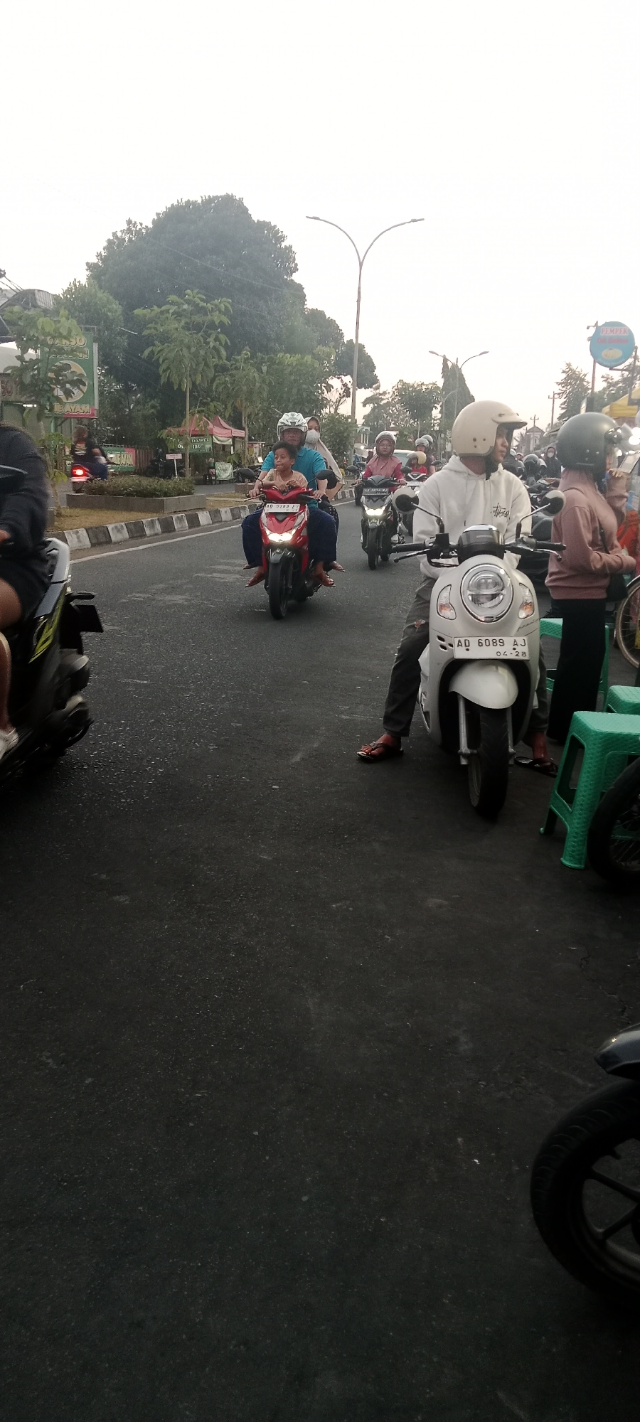 An outdoor, eye-level, medium shot captures a street scene in Ngemplak, Indonesia, likely during the late afternoon due to the overcast sky and diffused light. The focus is on a group of people on motorcycles.
In the foreground, a white scooter with a rider wearing a light-colored hooded sweatshirt and a beige helmet is prominently featured. The scooter's license plate reads "AD 6089 AJ 04-28." The rider's legs are visible, clad in dark pants and wearing flip-flops. To the right of the scooter, a green plastic stool is present.
Further down the street, a red scooter carries two individuals: a man in a blue shirt and white helmet, and a young child in the front wearing a tan shirt and white helmet. The scooter's license plate is partially obscured but shows "AD 5783 FJ."
Other motorcycles and riders are visible in the background, suggesting moderate traffic. One motorcycle on the left is partially out of frame, with its front wheel and part of the frame visible. Another motorcycle further back has a rider in a maroon outfit and a light-colored helmet.
The environment includes a paved road with visible white lane markings and a curb. Beyond the road, there are trees, some with lush green foliage, and a few structures, including what appears to be a food stall with a red tent. A street lamp with a distinctive curved design is visible in the mid-ground. A sign with text in Indonesian, possibly indicating a business, is also present on the left side of the image. The overall atmosphere suggests a typical day in a semi-urban or suburban area.