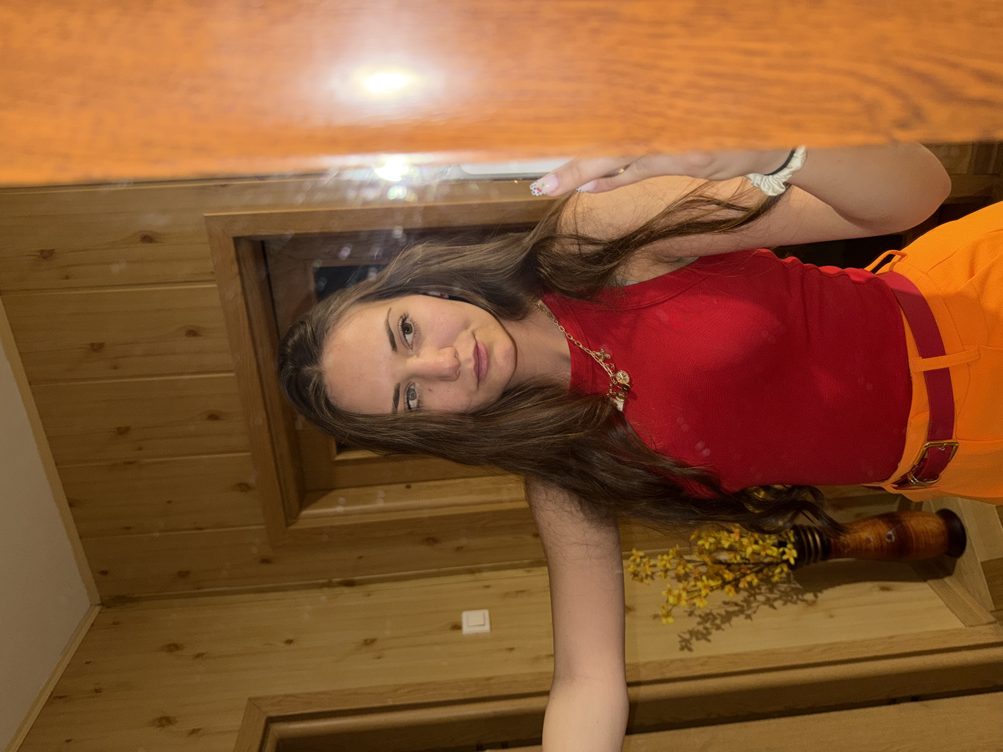The image is a selfie of a young woman with long brown hair wearing a red tank top, orange pants with a reddish-brown belt, and a gold necklace. She is positioned in what appears to be a wooden-paneled room. A wooden doorframe and wall are visible in the background, as well as a decorative vase with yellow flowers. The reflection in the mirror on top is a slightly blurred wood plank structure. There's no specific evidence to confirm it is Buhovo, Bulgaria.