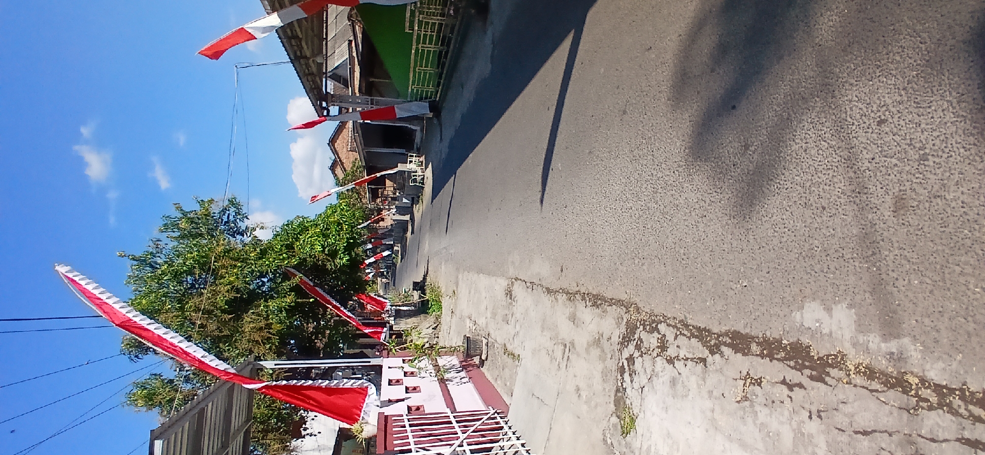 In the city of Pejaten, Indonesia, an asphalt road stretches into the distance, lined on both sides by houses and trees under a clear blue sky. Red and white flags, likely Indonesian flags, are prominently displayed along the street, tied to poles and draped from structures, suggesting a festive or patriotic occasion. The houses appear to be single-story structures with tiled roofs. The roadside on the left features a concrete sidewalk and a pink and white fence. The roadside on the right has a green fence and the eaves of several buildings. The sun casts long shadows across the road, indicating it is likely late afternoon. The scene depicts a quiet residential street decorated for a celebration.
