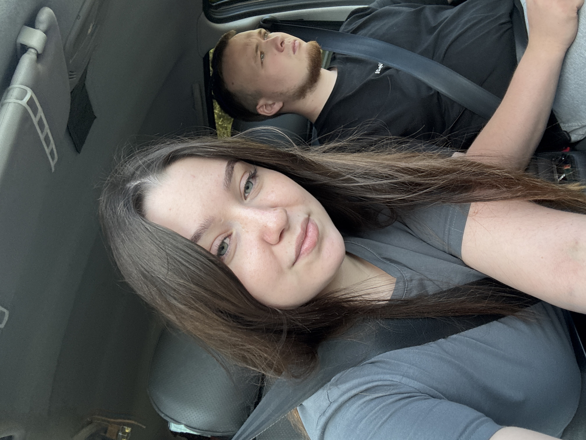 This is a medium shot, taken from inside a car, of a young couple. The woman is in the foreground, in the passenger seat, and is looking directly at the camera. She has long, dark brown hair with a streak of grey near her temple, and is wearing a grey t-shirt. Her eyes are green and she has a light dusting of freckles across her nose and cheeks. The man is in the driver's seat, to the right of the woman, and is looking out the side window. He has short, light brown hair and a beard, and is wearing a black t-shirt. Both individuals are wearing seatbelts. The interior of the car is visible, including the grey headliner and a sun visor. The scene appears to be set in a city, possibly Perm, Russia, given the visual context provided.