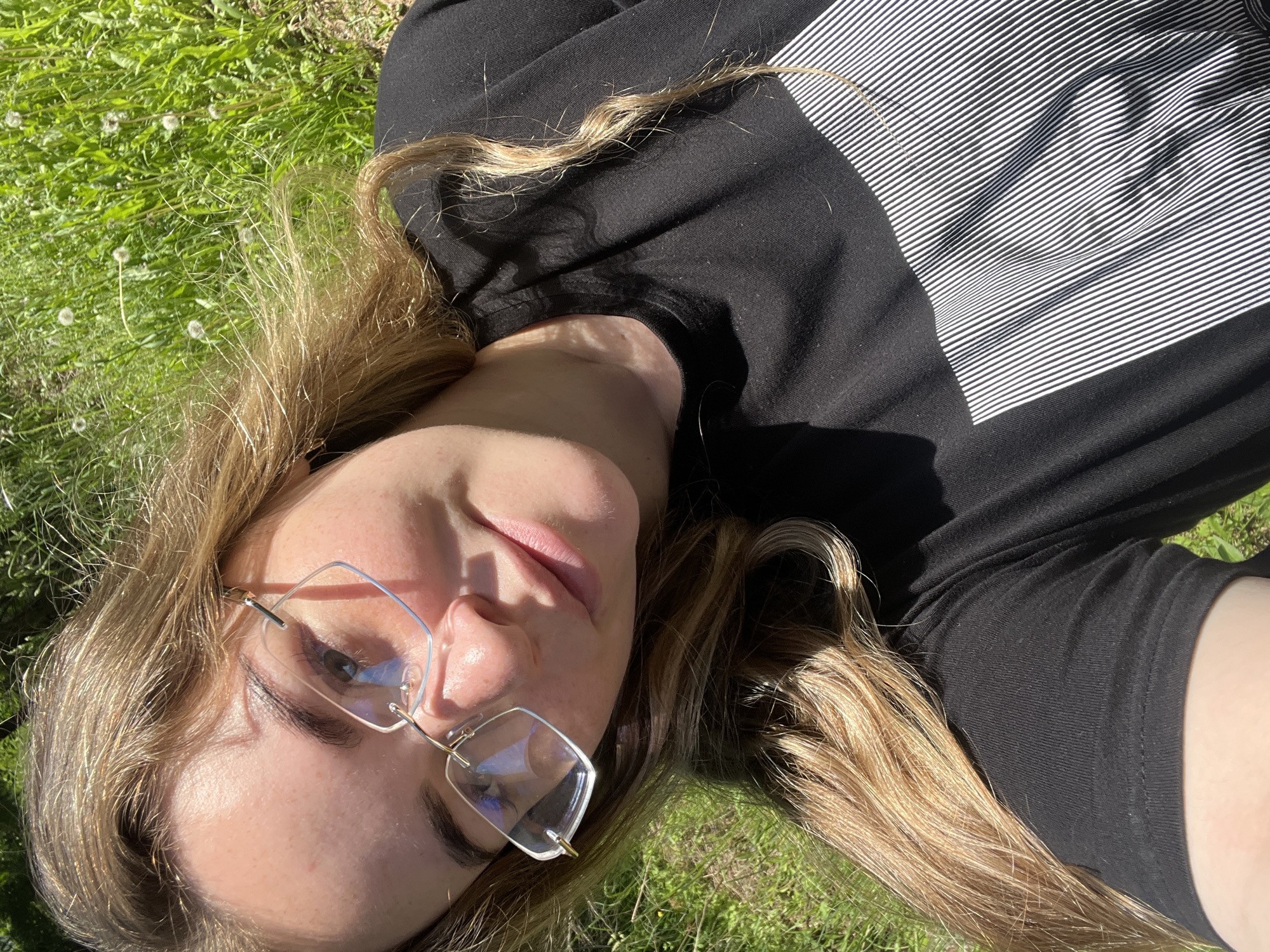 The image displays a young, light-skinned woman with long, light brown hair, lying on her back on green grass in Сосново, Russia. She is wearing rimless glasses with light-colored metal accents and an irregular hexagonal lens shape. Her eyes are visible behind the lenses, reflecting light. She is clad in a black short-sleeved t-shirt that features a rectangular design composed of thin white vertical stripes on its lower right portion. The woman is positioned with her head slightly tilted, looking directly towards the camera with a subtle smile. Sunlight strongly illuminates her face and hair, creating distinct shadows on the left side of her face, under her chin, and across her neck and shoulder. Her hair is spread out around her head, resting on the grass. The background consists of vibrant green grass intermingled with several white dandelion seed heads. The photograph is taken from a low angle, consistent with a selfie, showcasing the woman against the backdrop of the natural environment.