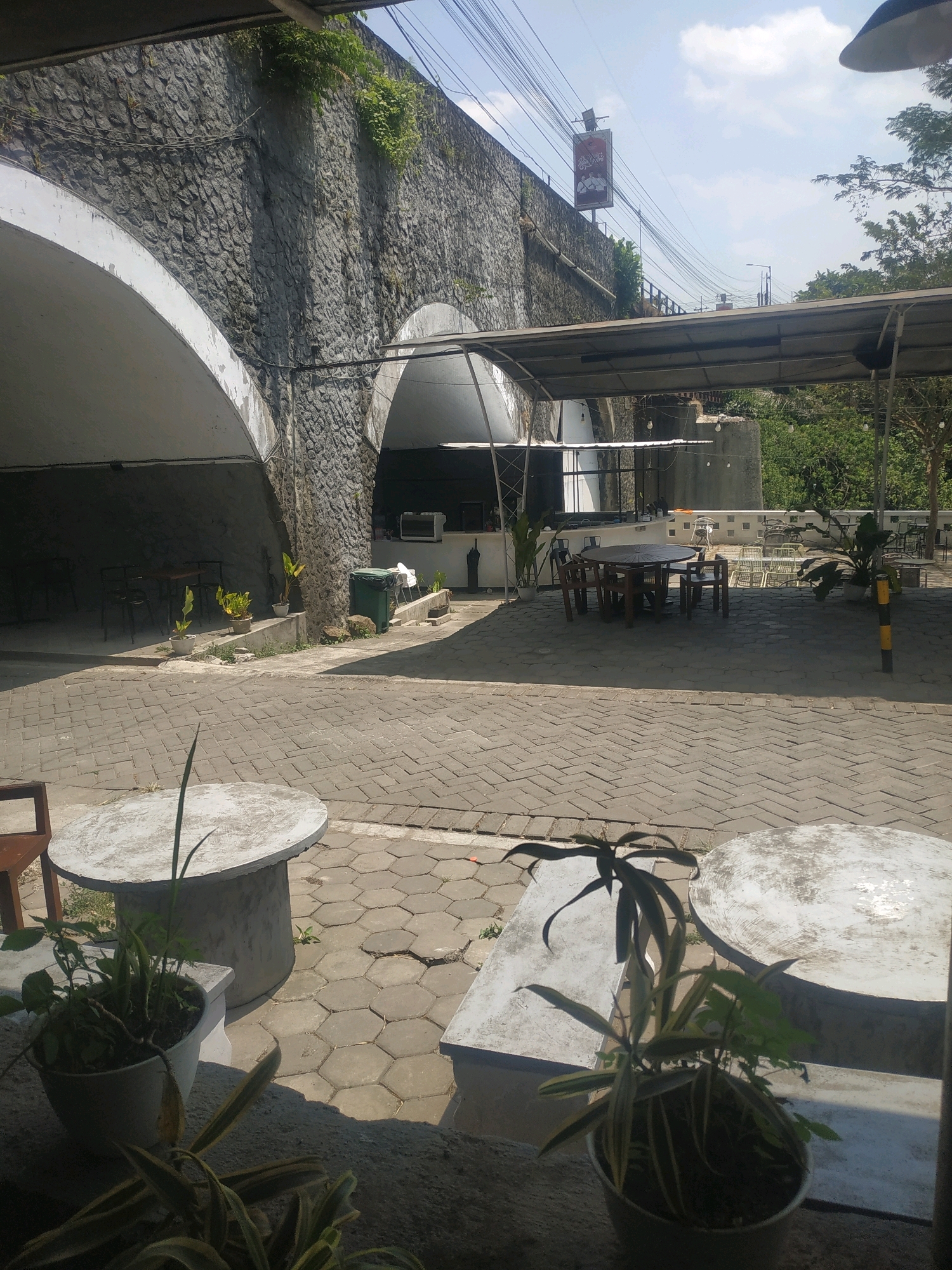 The image depicts an outdoor seating area with a stone-arched structure in the background. In the foreground, two round concrete tables and a potted plant are visible. The ground is paved with hexagonal concrete tiles. Further back, under a canopy, a wooden round table with chairs is set up, alongside other tables and chairs. A green trash can is situated near the stone structure. The scene is outdoors, with a bright sky and trees in the distance, suggesting a daytime setting. The location is identified as Patrang, Indonesia.