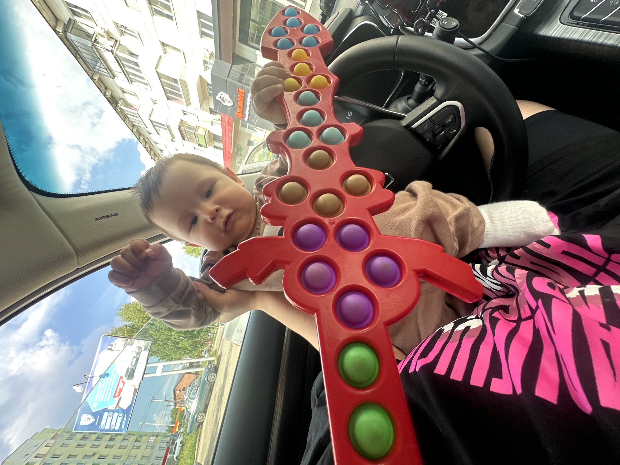 A low-angle, indoor shot from a car shows a baby holding a red toy sword with pop-it bubbles in varying shades of purple, blue, yellow, and green. The baby, positioned centrally, looks towards the camera with a neutral expression. The toy sword extends horizontally across the foreground, partially obscuring the baby's body. The background reveals the interior of a car, including a black steering wheel and dashboard. Through the car's windows, an urban outdoor environment is visible, featuring apartment buildings, trees, and a billboard with text in Russian. The sky is partly cloudy. The overall lighting suggests it is daytime.
