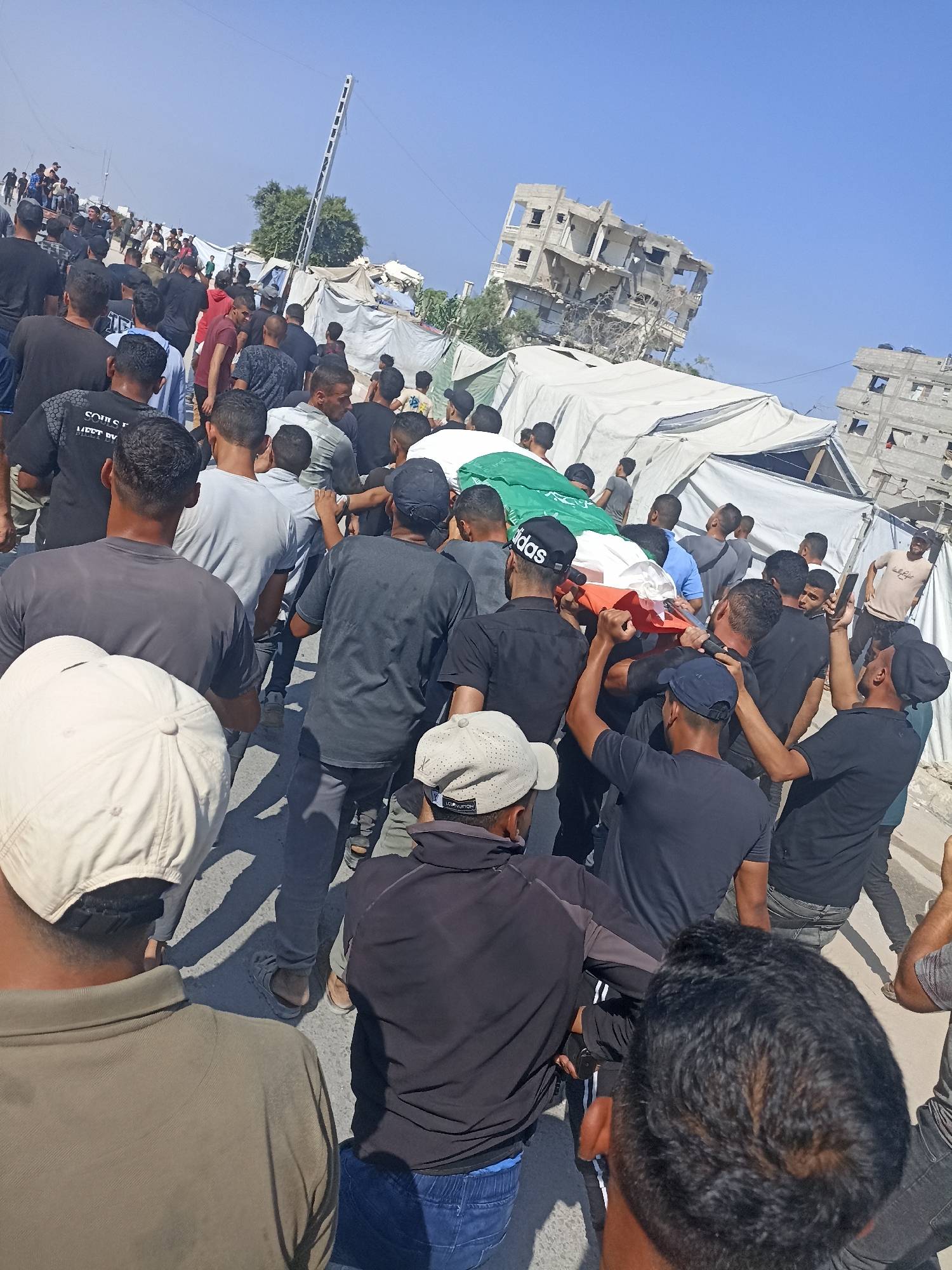 A procession of men carries a flag-draped coffin through a street lined with tents and damaged buildings in Gaza City, Palestinian Territories. The coffin is wrapped in a flag that appears to be the Palestinian flag. The men are mostly wearing dark clothing and some have head coverings. The background shows destroyed buildings, suggesting a conflict zone. The sky is clear and blue.