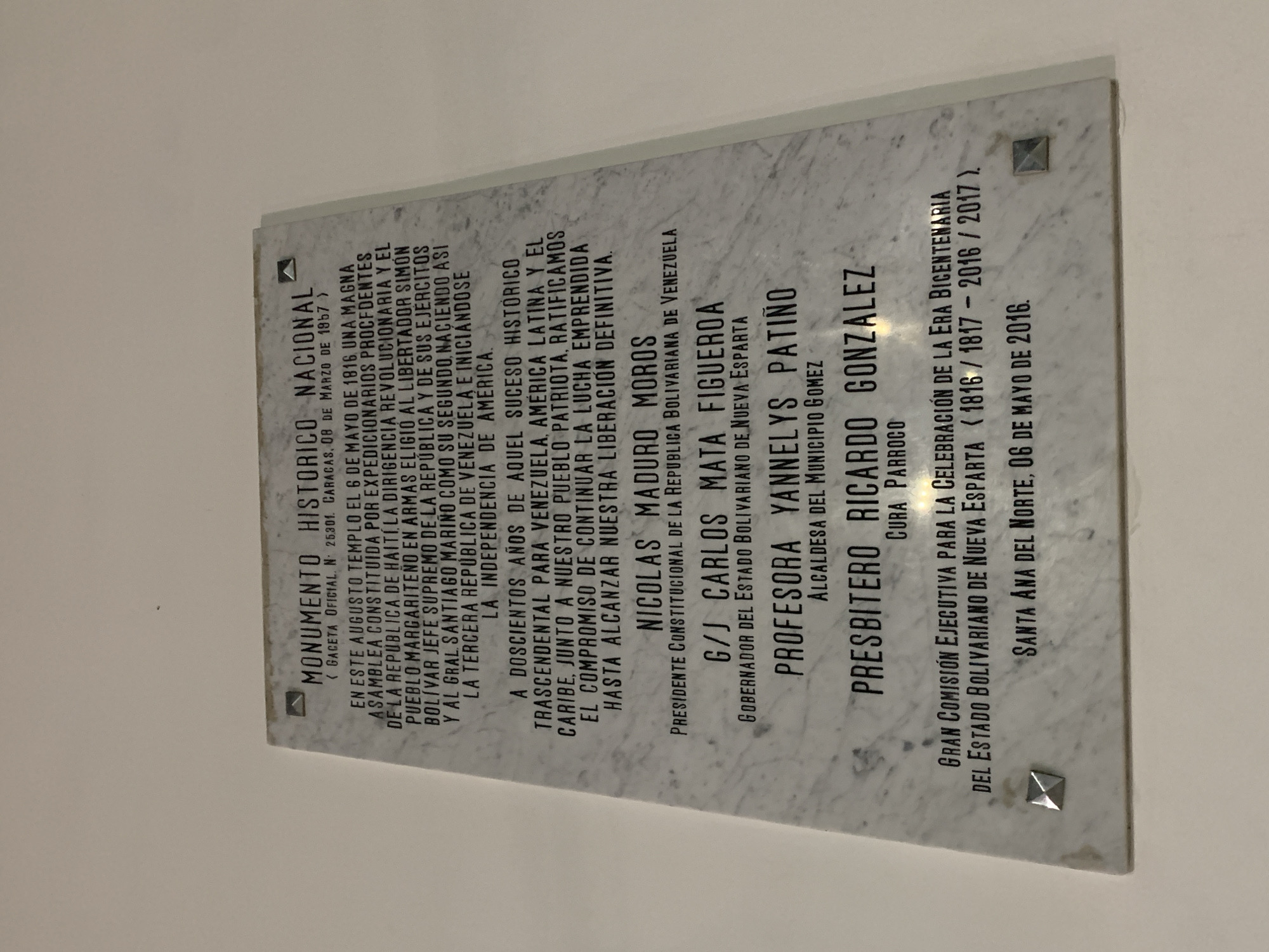 A rectangular, light-colored marble plaque is prominently displayed on a plain, light-colored wall. Four metallic, pyramid-shaped fasteners secure the plaque at each corner.
The plaque is titled "MONUMENTO HISTÓRICO NACIONAL" (National Historical Monument) and references "GACETA OFICIAL. N. 25.301. CARACAS, 08 DE MARZO DE 1967." The main text commemorates a significant historical event: on May 6, 1816, in this "august temple," an assembly formed by expeditionaries from the Republic of Haiti, revolutionary leadership, and the armed people of Margarita, elected Simón Bolívar as Supreme Chief of the Republic and its armies, and General Santiago Mariño as his second. This pivotal event initiated the Third Republic of Venezuela and its independence.
The plaque further states that 200 years after this transcendental historical event for Venezuela, Latin America, and the Caribbean, the patriotic people ratify their commitment to continue the struggle until definitive liberation is achieved.
It lists the following officials involved in the commemoration:
- NICOLÁS MADURO MOROS, Constitutional President of the Bolivarian Republic of Venezuela.
- G/J CARLOS MATA FIGUEROA, Governor of the Bolivarian State of Nueva Esparta.
- PROFESORA YANNELYS PATIÑO, Mayor of Gomez Municipality.
- PRESBÍTERO RICARDO GONZÁLEZ, Parish Priest.
The plaque also acknowledges the "GRAN COMISIÓN EJECUTIVA PARA LA CELEBRACIÓN DE LA ERA BICENTENARIA DEL ESTADO BOLIVARIANO DE NUEVA ESPARTA (1816 / 1817 - 2016 / 2017)." The commemorative plaque itself is dated "SANTA ANA DEL NORTE, 06 DE MAYO DE 2016." The setting appears to be indoors or a covered area given the uniform background and lighting.