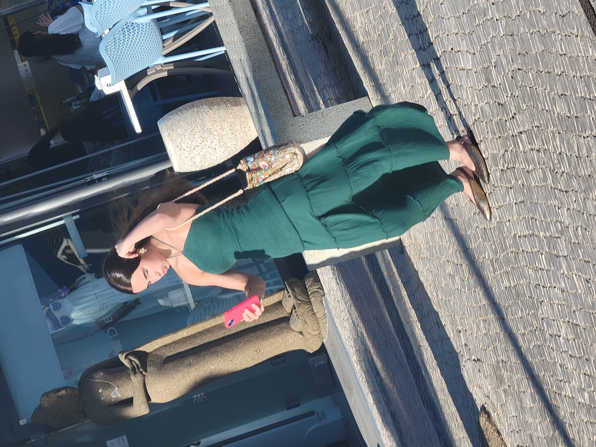 A person is seated outdoors, holding a pink smartphone. They are wearing a long, emerald green dress and metallic gold shoes. A small, embroidered purse with a rope strap hangs from their shoulder. The ground beneath them is paved with textured gray tiles that cast long shadows, suggesting it is late afternoon. In the background, there are light blue chairs and a white woven object. To the left, a glass surface reflects other people and a statue-like object. The setting appears to be a public area, possibly a promenade or terrace, given the seating and the presence of a statue. The sunlight is bright, indicating fair weather. The person's posture and the way they are holding their phone suggests they might be taking a photo or checking a message.