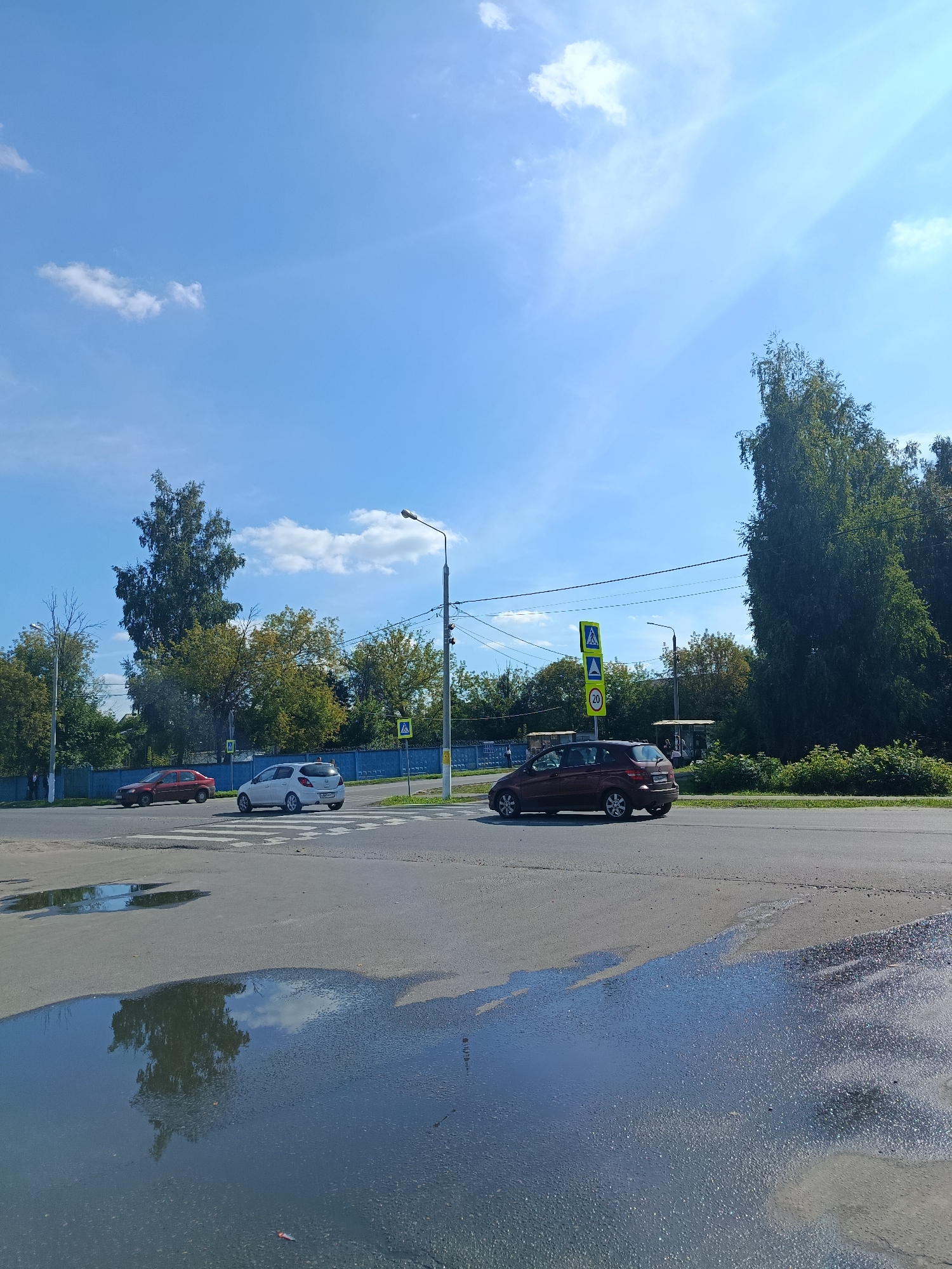 The image shows a street scene in Orekhovo-Zuyevo, Russia, on a clear, sunny day with scattered clouds. In the foreground, a large puddle reflects the sky and trees. The middle ground features a road with a pedestrian crossing. Several cars are visible: a red hatchback, a white hatchback, and a dark red SUV. The white hatchback is positioned on the pedestrian crossing, and the red hatchback is further down the road. The dark red SUV is moving away from the camera on the right side of the road. Traffic signs are present, including a pedestrian crossing sign and a speed limit sign (20 km/h). Trees and foliage line the background, with a blue fence visible to the left. A bus stop shelter with people can be seen in the distance on the right. Streetlights are also visible. The overall impression is a typical urban street scene with moderate traffic.