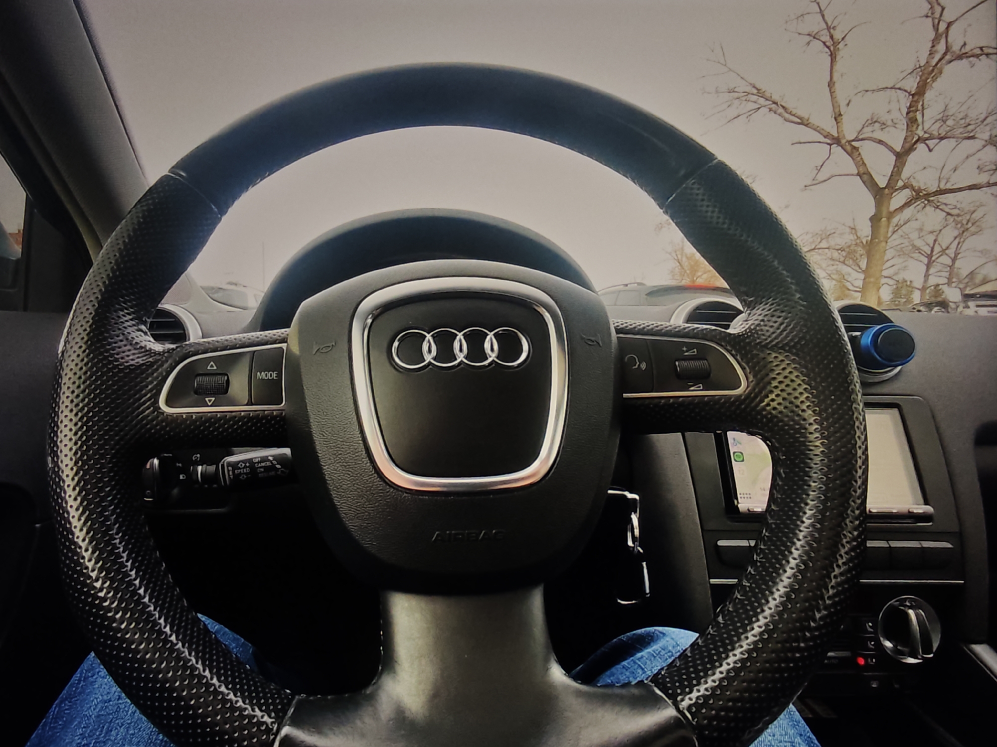 The image is a close-up, first-person perspective from inside an Audi car, showing the steering wheel and dashboard. The steering wheel is black with a textured grip and features the Audi logo in the center. Buttons for cruise control and other functions are visible on the steering wheel. A navigation screen is also visible to the right of the steering wheel. The driver's legs, clad in blue jeans, are visible at the bottom of the frame. Outside the car, through the windshield, bare trees and an overcast sky can be seen, suggesting a winter or early spring setting. There are no visible indicators of the city of Calgary, Canada in the image.