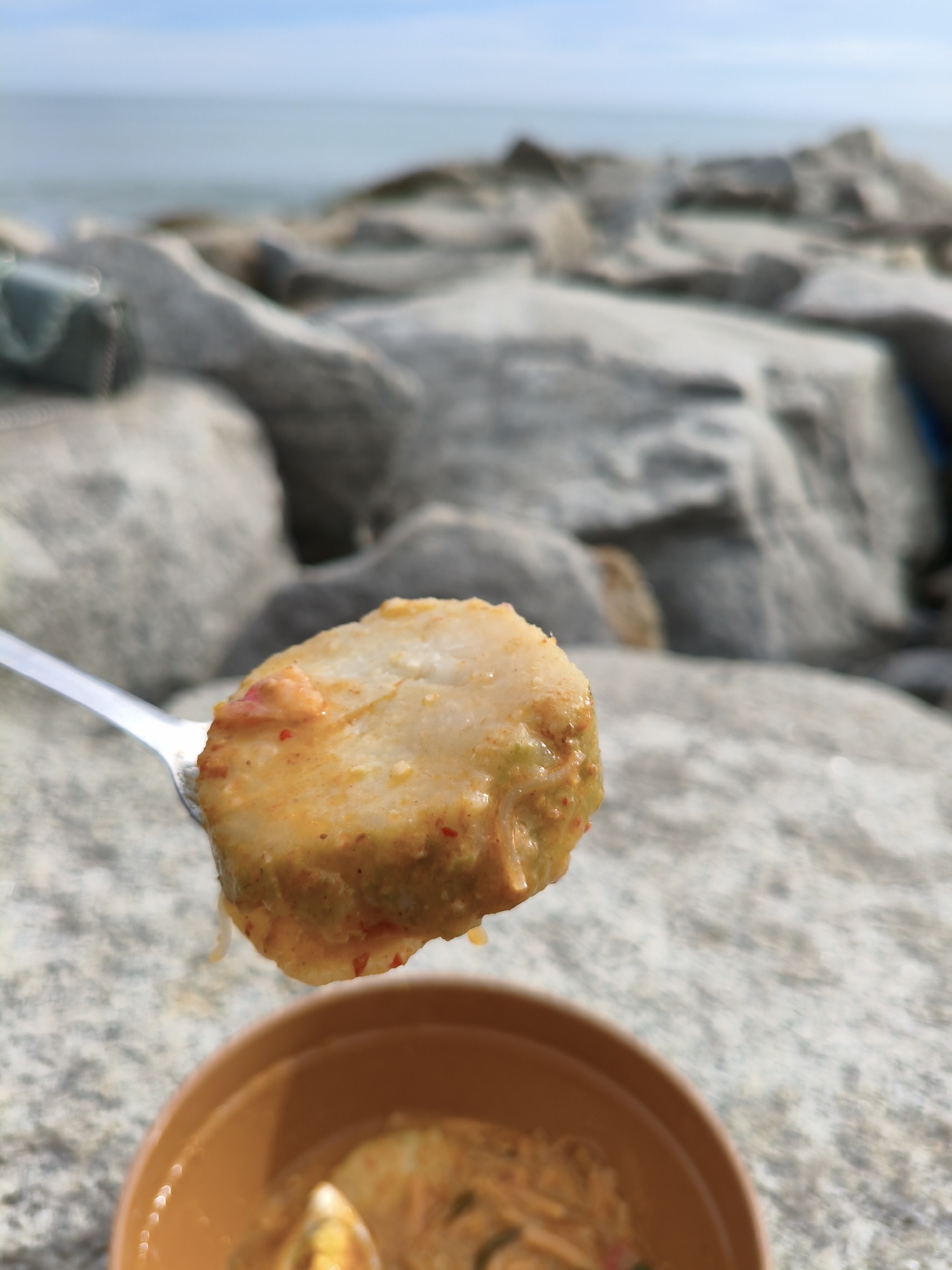 A close-up, shallow depth-of-field photograph shows a spoon holding a piece of food with a sauce, poised above a brown bowl. The food on the spoon is rectangular and appears to be a savory dish, possibly noodles or rice cake, coated in a yellowish-orange sauce with visible red flecks and some green elements. The background is out of focus, revealing large, grey rocks that suggest a coastal environment. Beyond the rocks, a blurred body of water and a hazy sky are visible, indicating a seaside location. The overall impression is of a meal being enjoyed outdoors. The scene is likely on a breakwater or rocky shore in Lhokseumawe, Indonesia, a city known for its coastal areas.